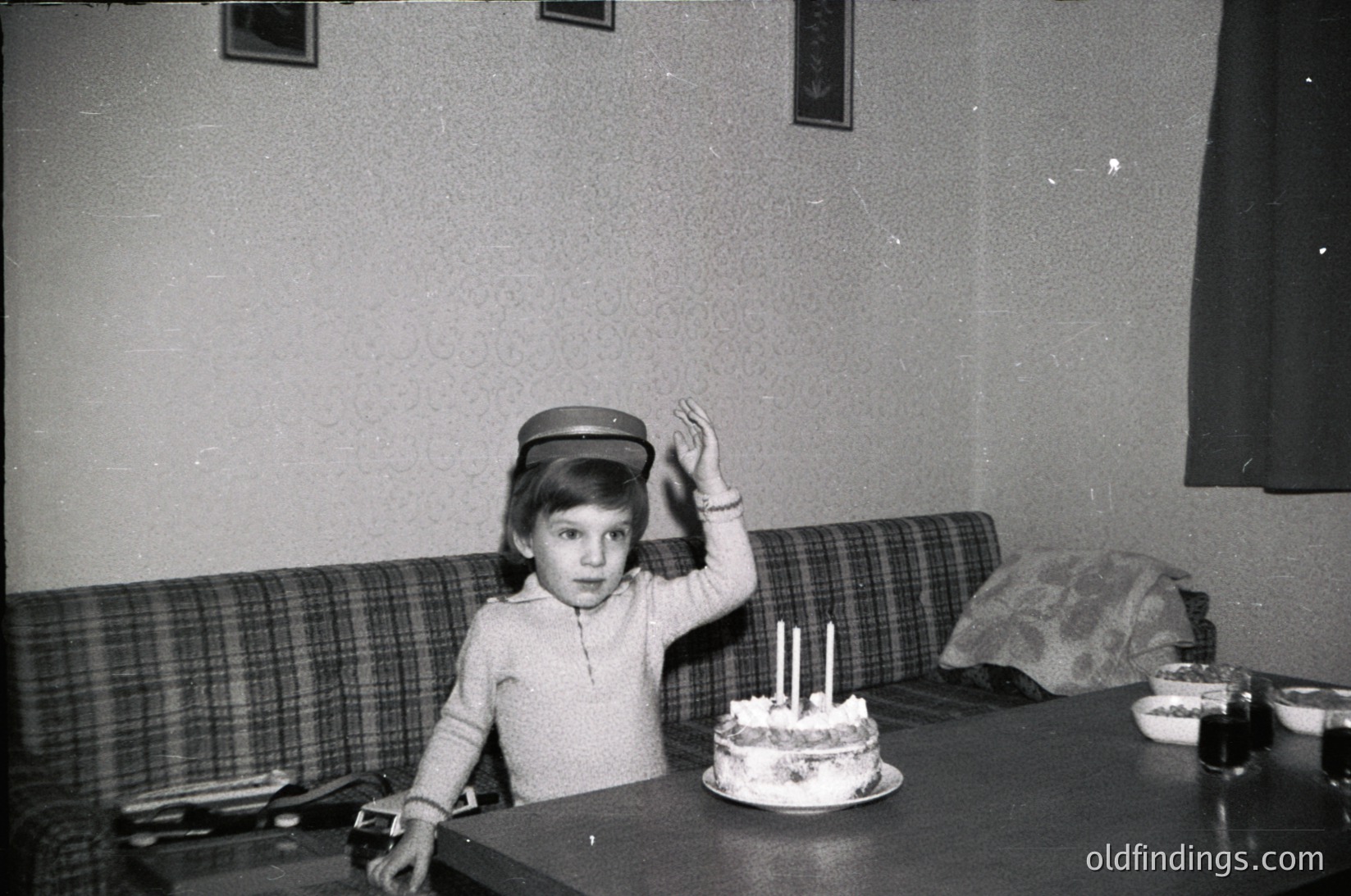 Child in 1960s-70s indoor setting, seated at a wooden table with a small birthday cake (three lit candles) and floral pattern. Wears a knitted sweater, wristwatch, and a flat cap. Plaid sofa and patterned pillow in background. Classic mid-century home decor.