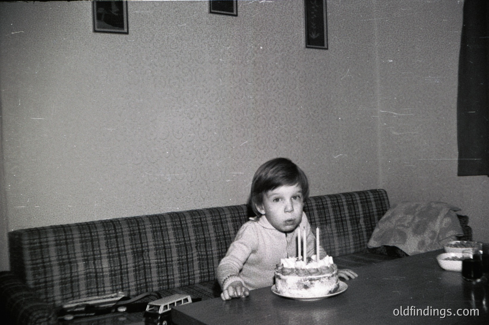 Vintage black-and-white photo of a young child seated at a table, blowing out candles on a round birthday cake. Mid-century interior with plaid sofa, wall-mounted stove, and framed art. Likely late 20th century domestic setting.