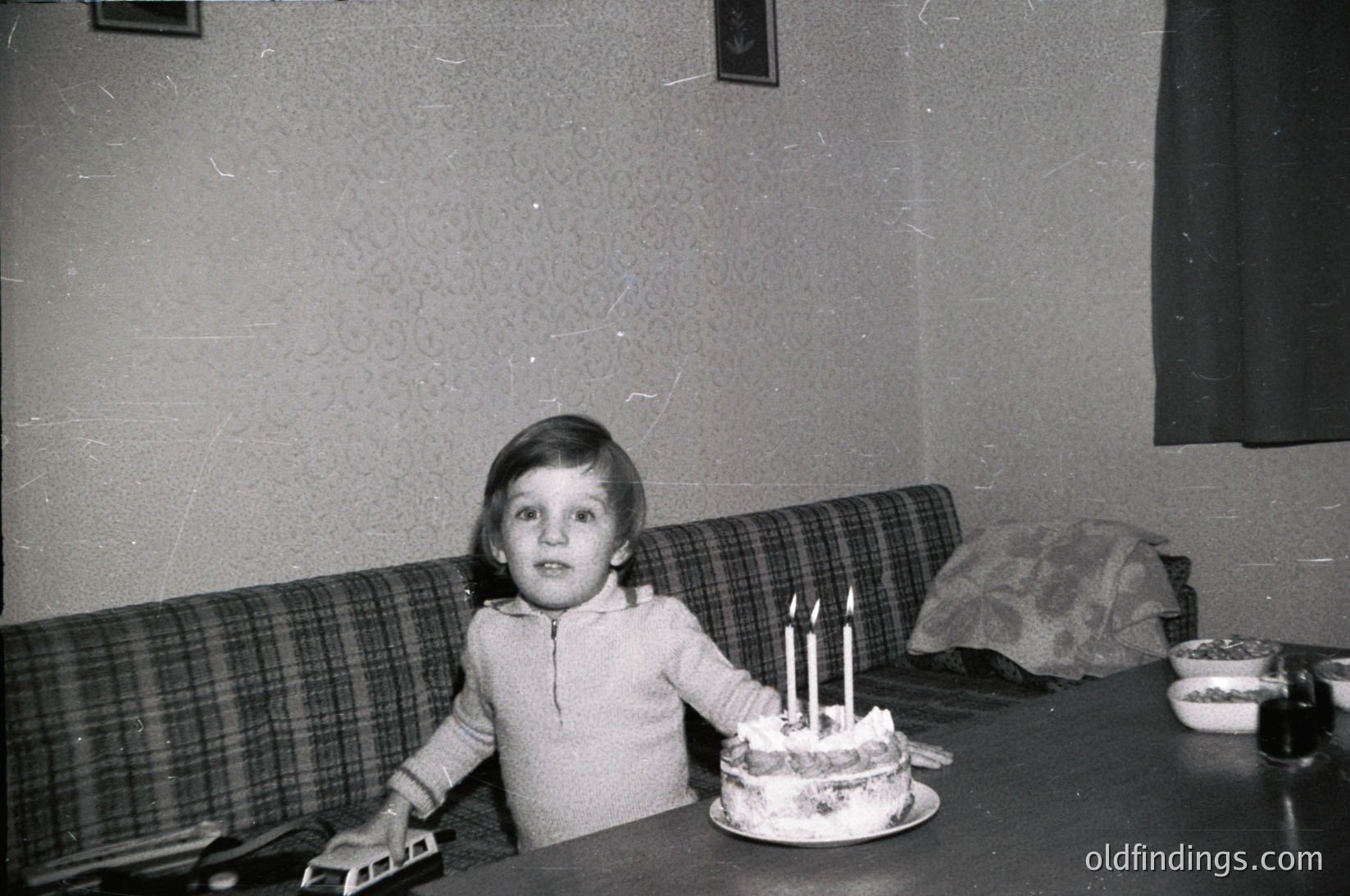 Vintage black-and-white photo of a child seated at a wooden table, blowing out three lit candles on a round cake. Plaid sofa and floral wallpaper suggest mid-20th century domestic setting.