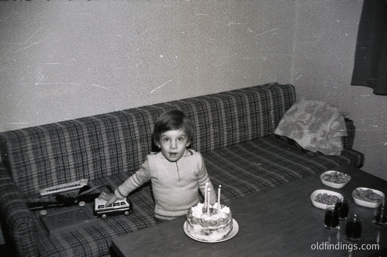 Black-and-white snapshot of a child seated on a plaid sofa, holding a toy vehicle while staring at a small birthday cake with lit candles on a wooden table. Mid-20th century domestic setting, likely 1960s–1970s.