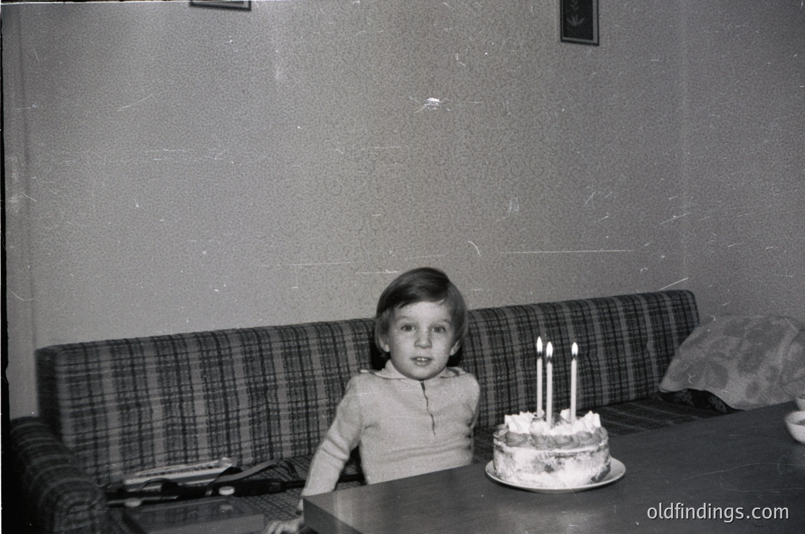 Vintage black-and-white photo of a child seated at a table with a three-candle birthday cake. Plaid sofa and worn wallpaper suggest mid-20th century domestic setting. Simple, nostalgic celebration.