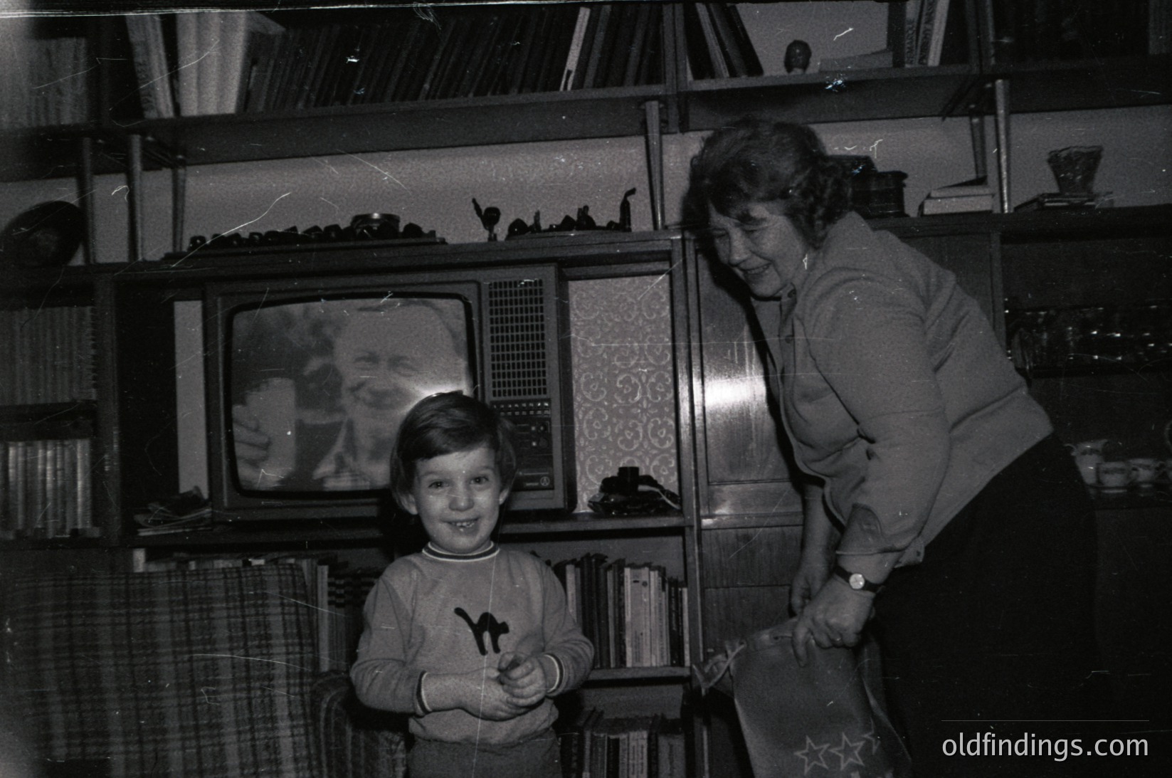 Mid-20th century living room scene: a woman in a patterned blouse and a boy in a sweater pose beside a vintage CRT TV (1960s-70s). Bookshelves filled with books and decorative items frame the TV. Warm lighting highlights the intimate moment.