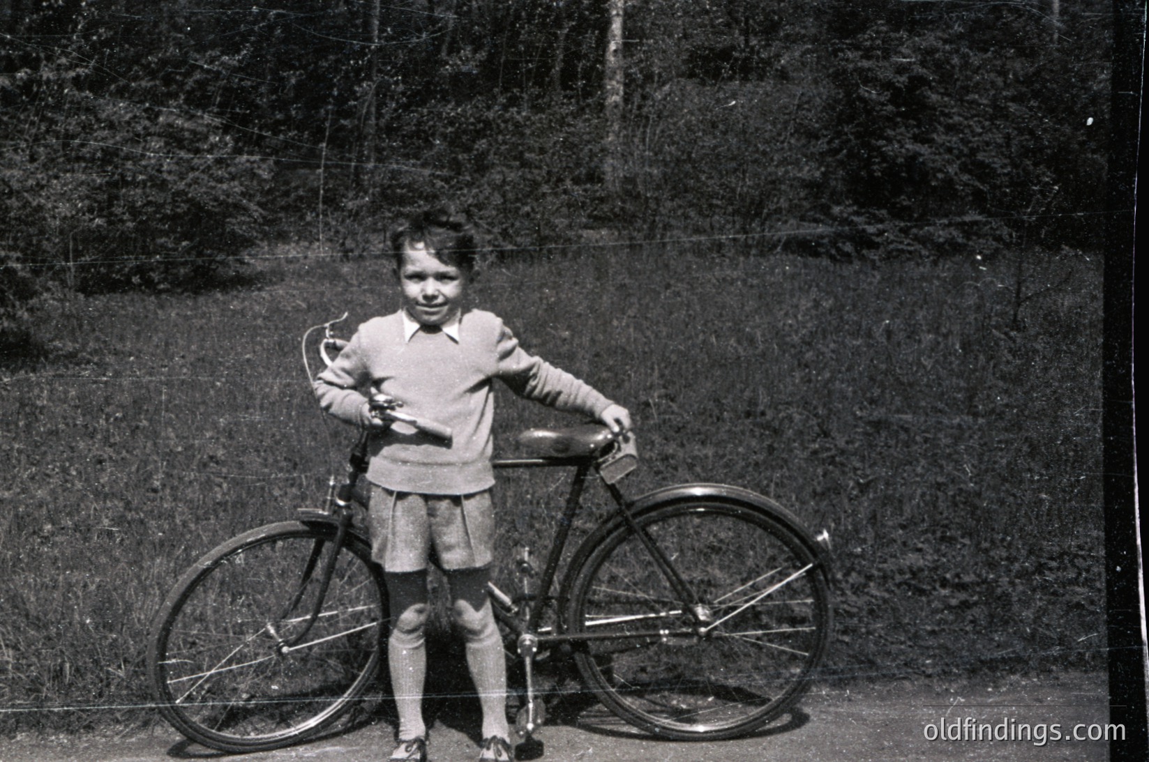 Vintage black-and-white photo of a young girl (approx. 6-8 years) posing beside a classic 10-speed bicycle in an outdoor setting. She wears a short-sleeve sweater, pleated skirt, and knee-high socks. The background shows dense foliage and a dirt path, suggesting a rural or suburban park. Likely mid-20th century (1950s–1970s).