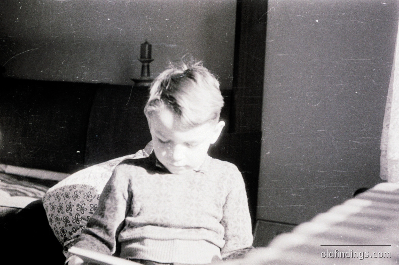 Mid-20th century black-and-white portrait of a young child seated indoors, wearing a patterned sweater and dark trousers. The blurred background suggests a modest, possibly rural setting with a wall-mounted lantern.