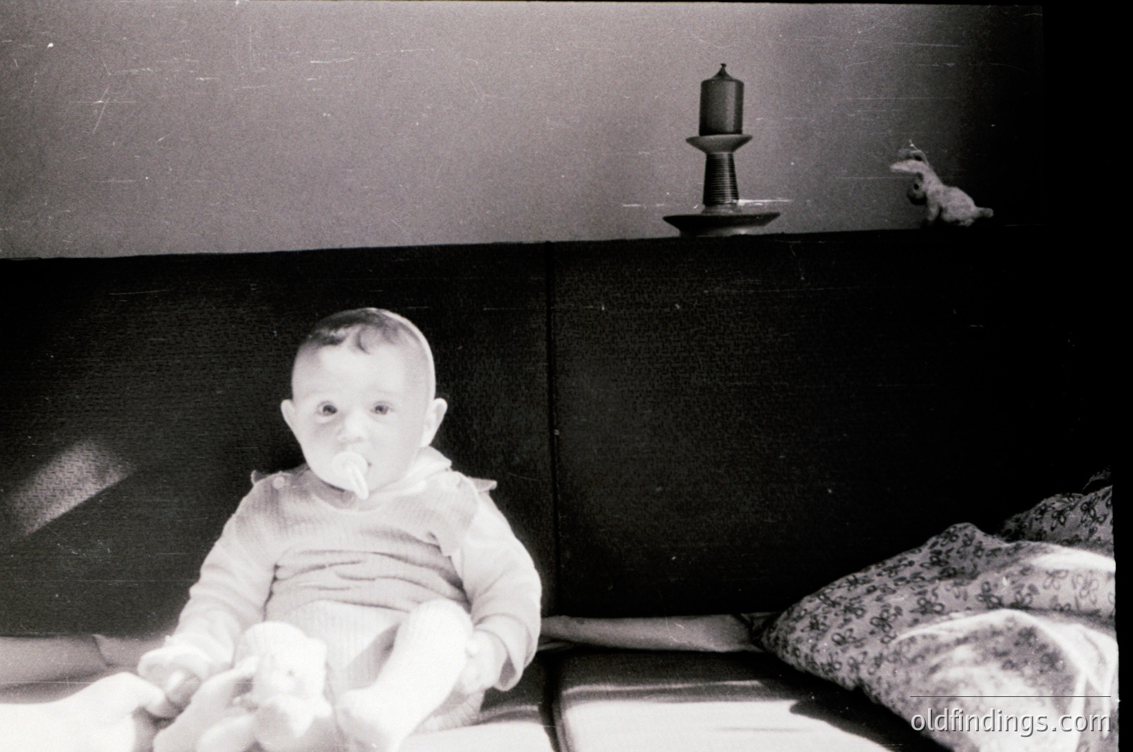 Vintage black-and-white photo of an infant seated on a bed, holding a toy. Simple wooden nightstand with a candle holder and stuffed animal. Mid-20th century domestic interior, likely 1950s–1960s.