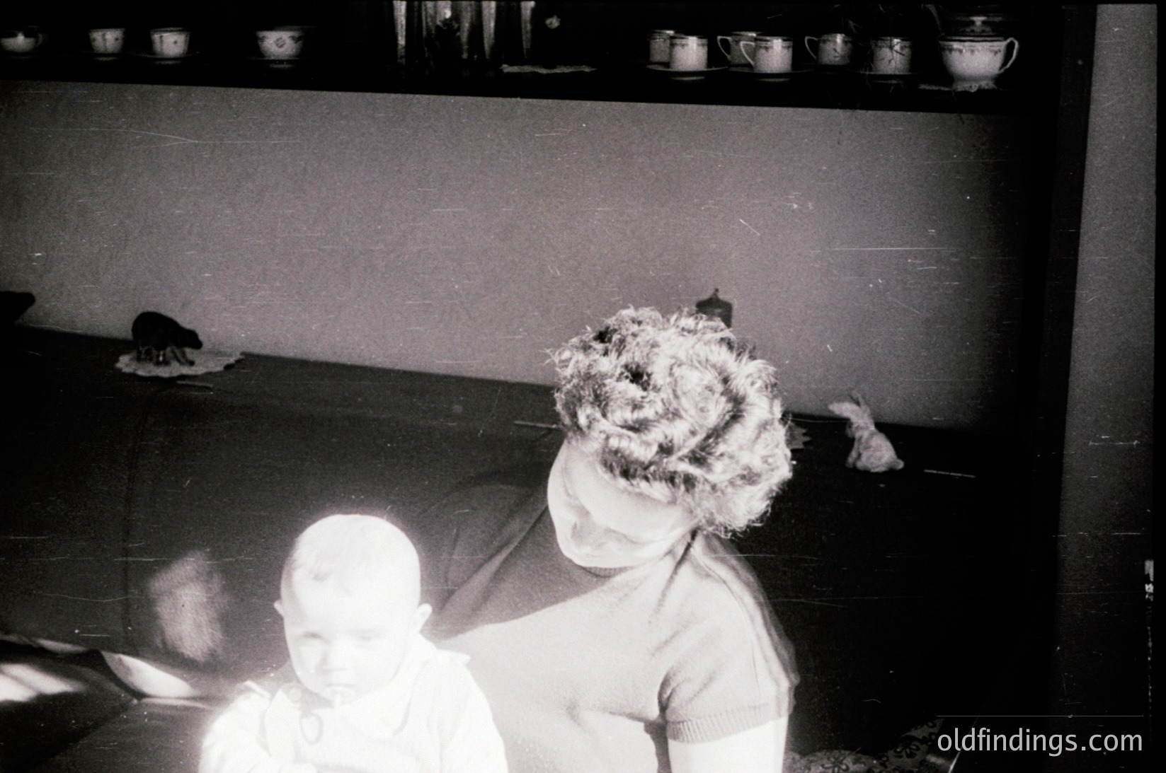 Vintage black-and-white photo of two children playing on a cluttered floor with a shelf of white ceramic mugs in background. Child on right wears a voluminous, curly hairstyle. Blurred motion suggests candid, spontaneous moment. Likely mid-20th century domestic setting.