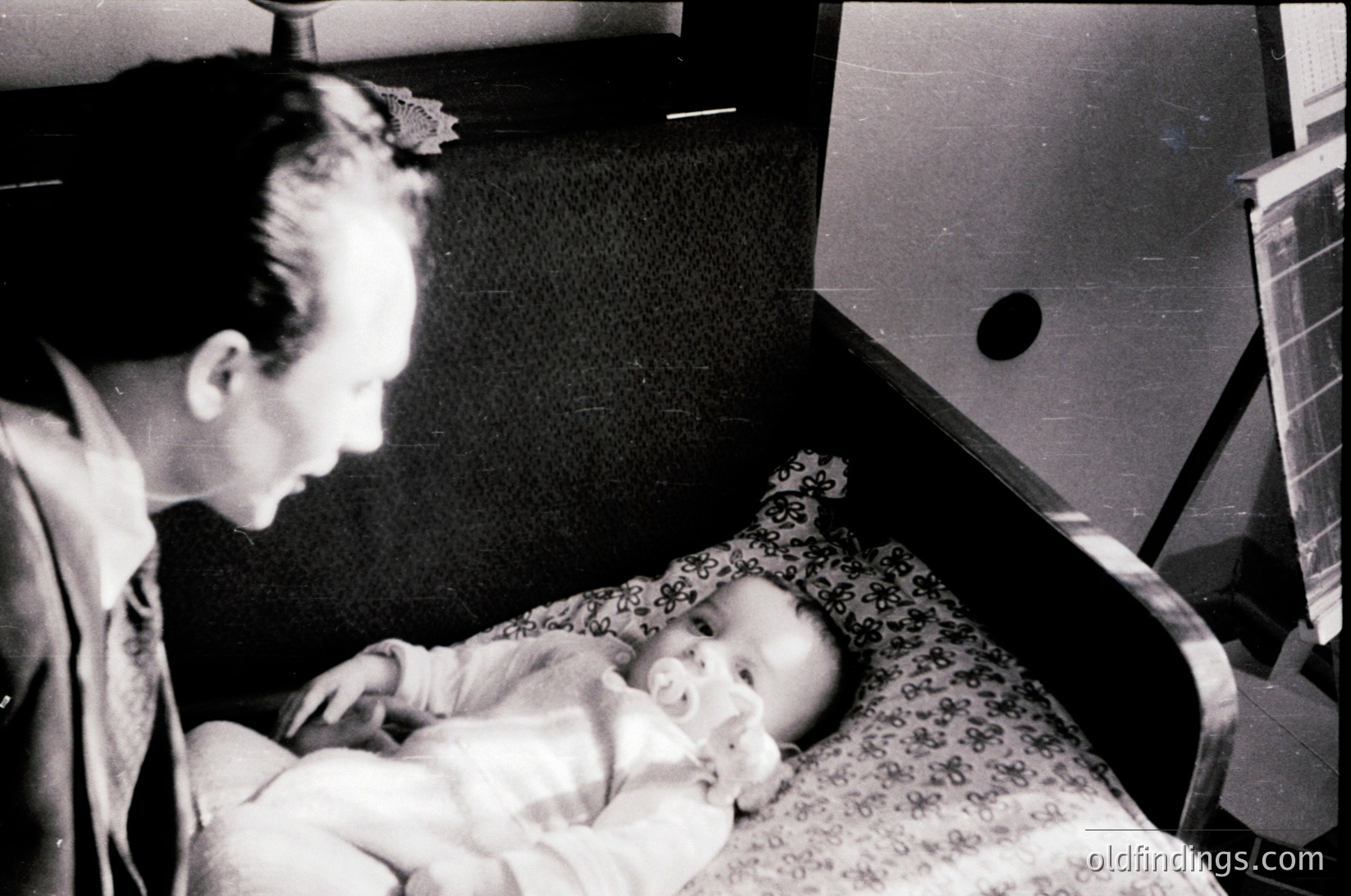 A man in mid-20th-century attire cradles a sleeping infant in a hospital bassinet, likely post-WWII or Cold War era. Floral-patterned bedding and a vintage oxygen tank in the background suggest a clinical setting.