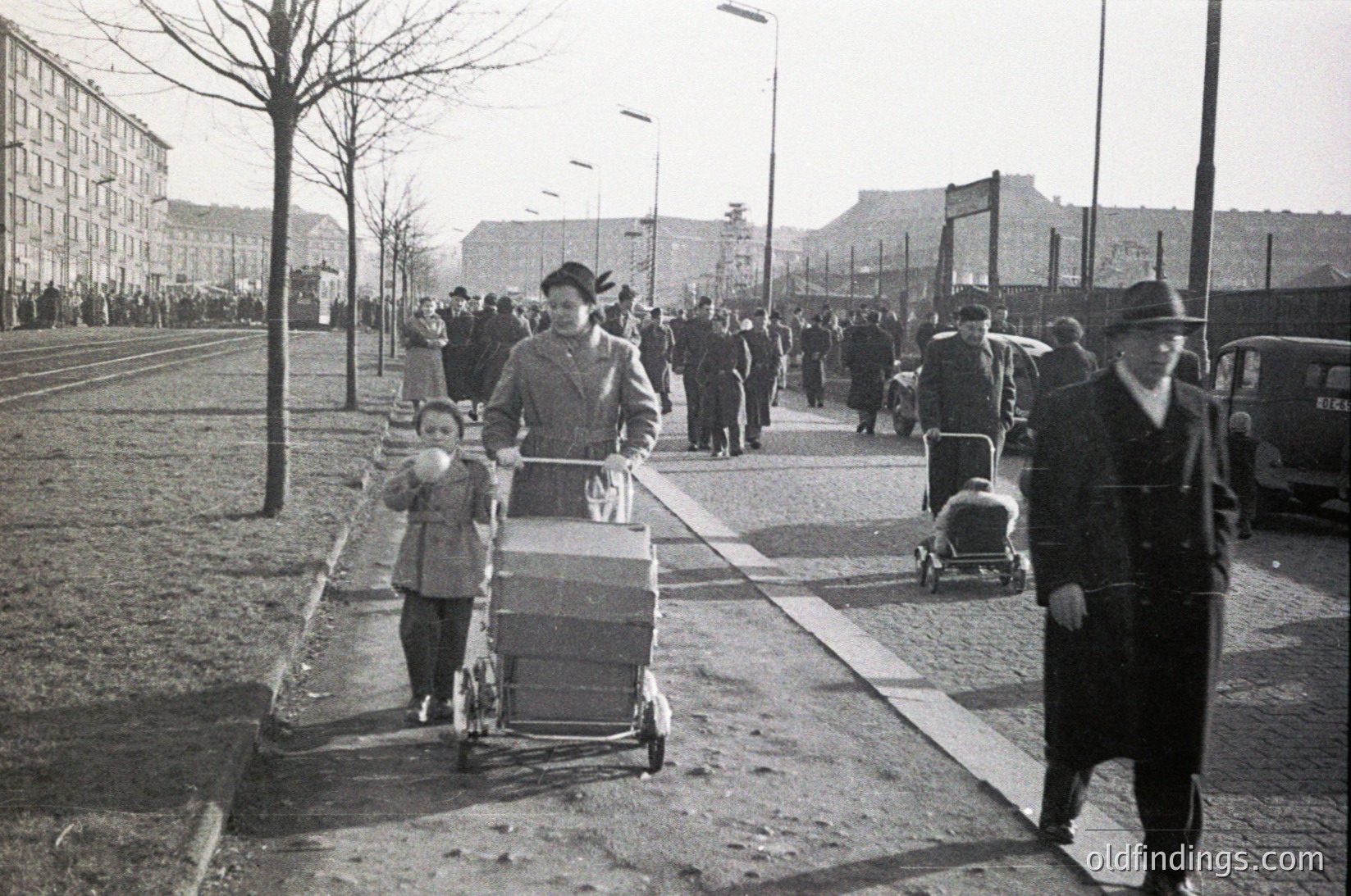 Mid-20th century urban street scene with families pushing strollers, likely post-WWII. Concrete sidewalks, bare trees, and a fence with barbed wire in background suggest restricted or divided area.