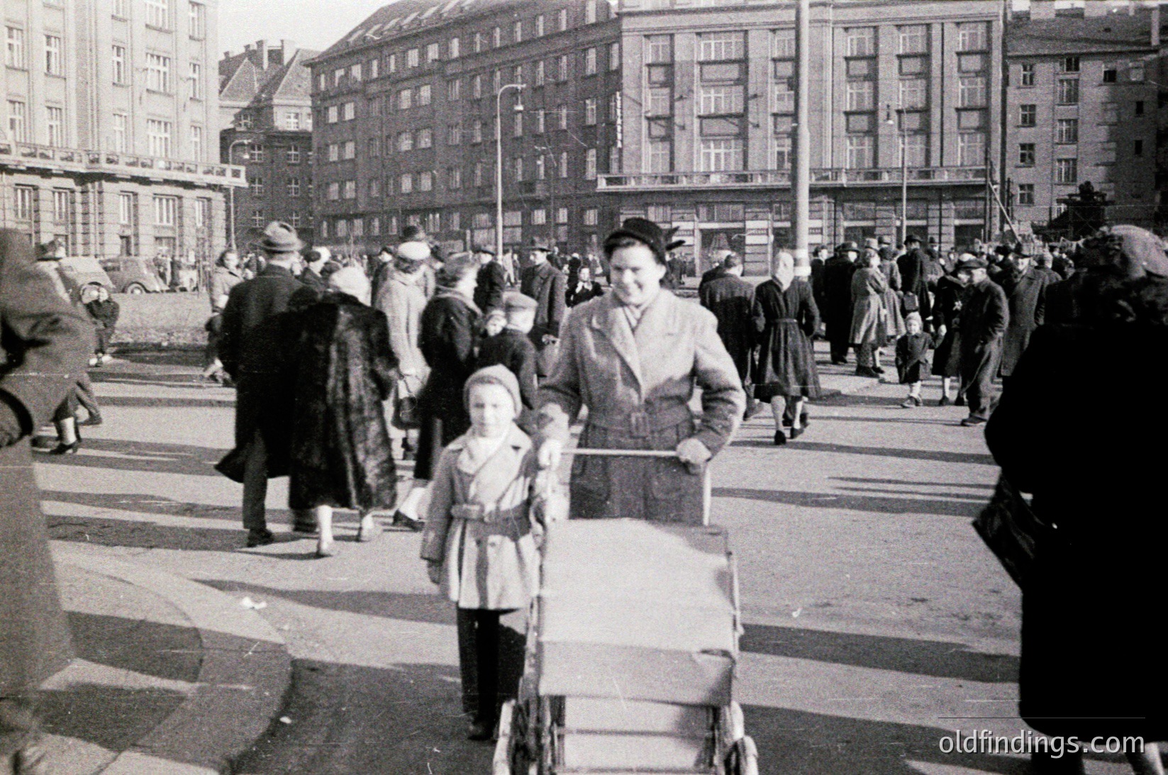 Mid-20th century urban street scene with Soviet-style architecture. Crowded plaza featuring a woman pushing a baby stroller, flanked by bundled children in winter coats. Multi-story concrete buildings with uniform windows dominate the background. Likely Eastern Bloc, 1950s–1960s.