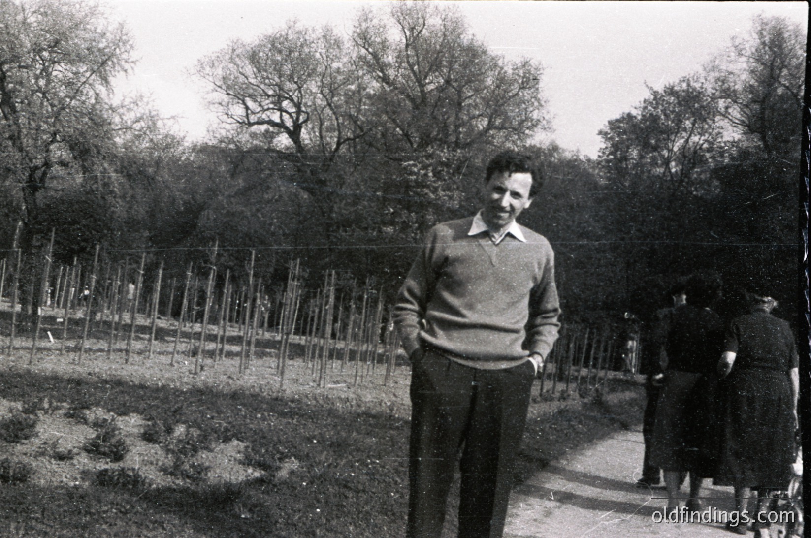 Black-and-white photo of a man in a sweater and trousers standing in a fenced garden area, likely a public park or communal garden. Bare trees and leafless shrubs suggest late autumn/winter. Fence posts and pathways indicate structured landscaping. Mid-20th century attire and setting suggest or European urban environment.
