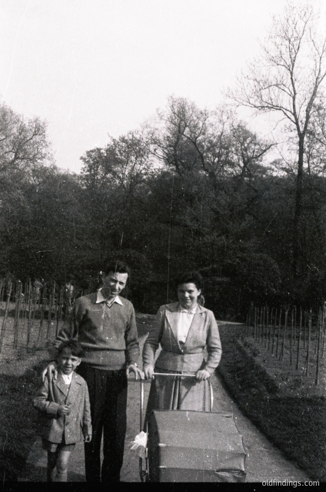 Black-and-white photo of a family (adults and child) posing outdoors on a paved path beside a fence and leafless trees, mid-20th century. Adults wear long-sleeve button-ups, cardigans, and skirts/pants; child wears a short-sleeve shirt and trousers. The woman holds a cane or walking stick.