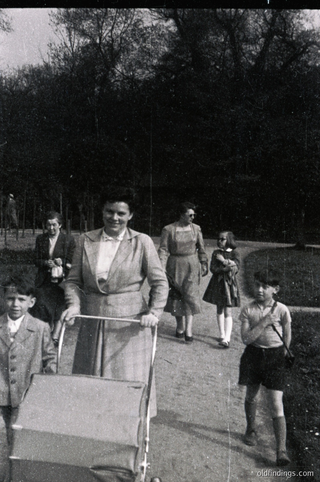 A mid-20th century family stroll in a park, likely 1950s–1960s. A woman in a structured blazer pushes a baby stroller, flanked by two young boys in short pants and long-sleeve shirts. In the background, two adults and a child walk toward a wooded area. Classic mid-century fashion and outdoor recreation capture everyday life.