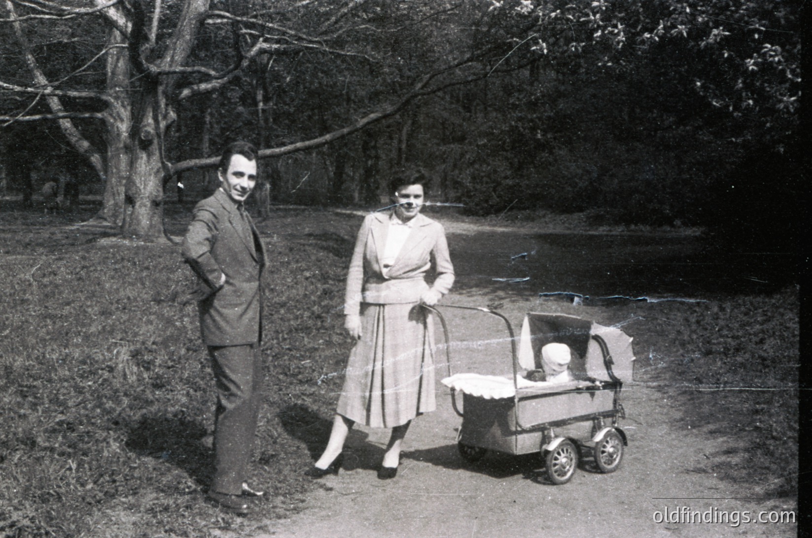 Mid-20th century couple posing outdoors with vintage stroller, likely 1950s–1960s. Man in suit, woman in knee-length dress with round sunglasses. Wooded path and natural lighting suggest a suburban or park setting.