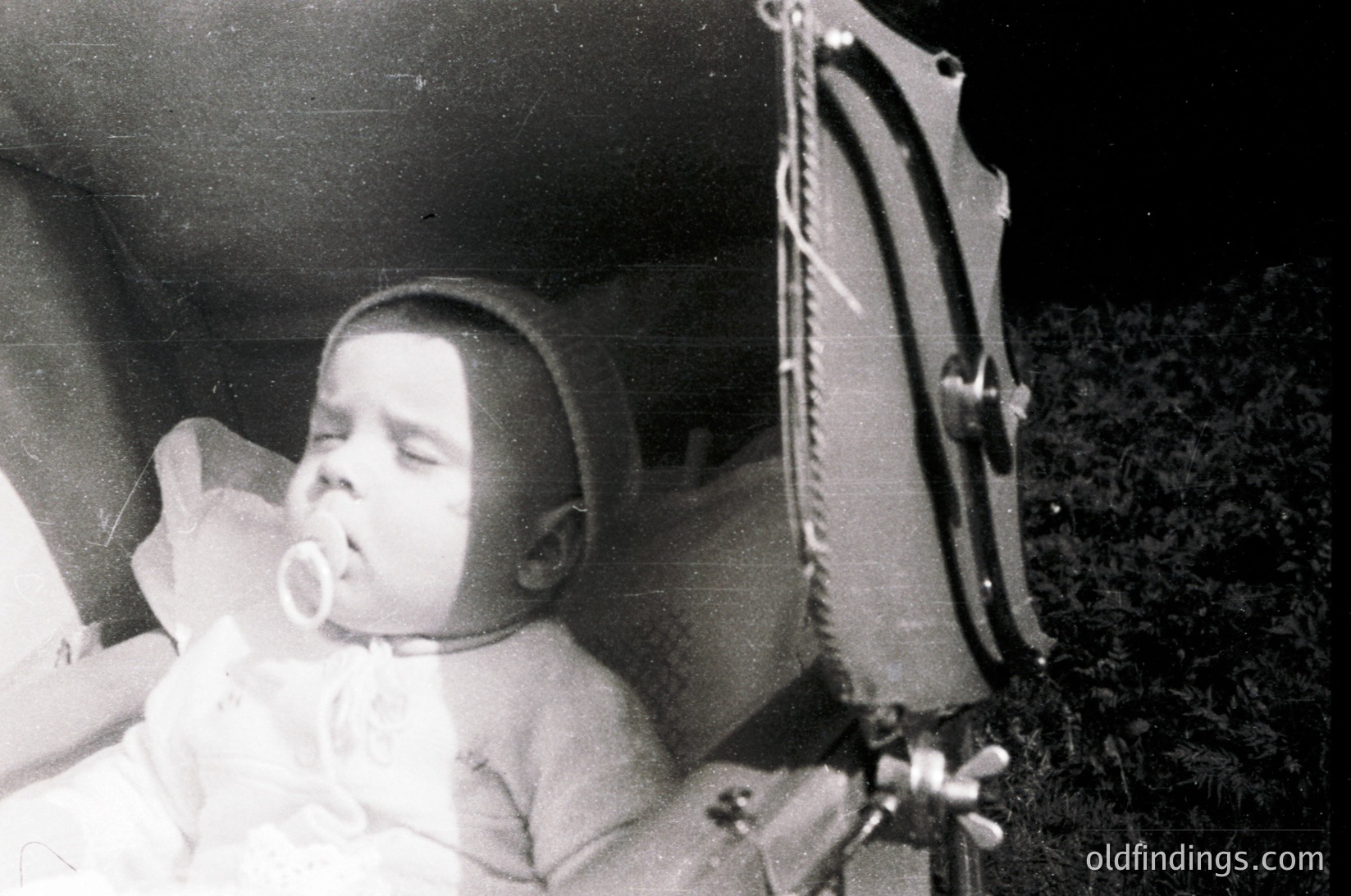 Vintage black-and-white photo of an infant sleeping in a crib with a pacifier, dressed in a simple onesie. Partial view of a wooden rocking chair frame on right. Soft lighting suggests indoor domestic setting, likely mid-20th century.