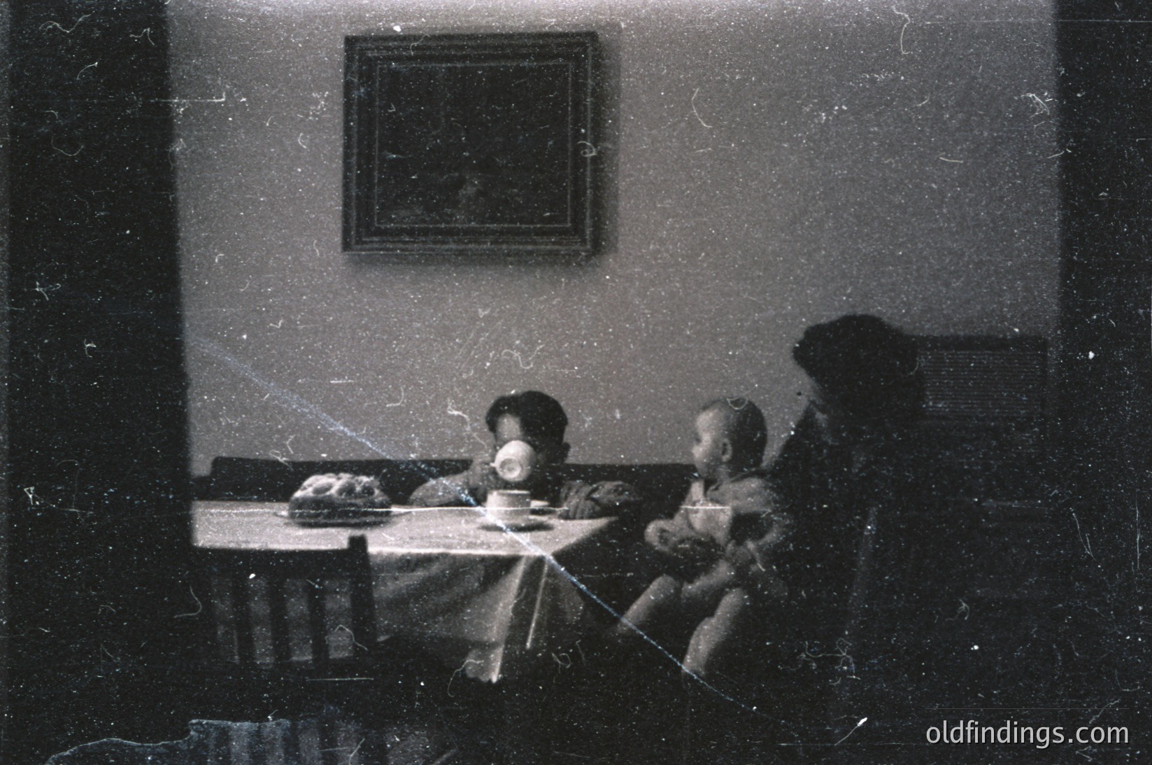 Vintage sepia-toned indoor scene featuring three seated figures at a table with a cake. Adults and child in formal attire, likely early-to-mid 20th century. Plain framed portrait above table, minimalist decor.