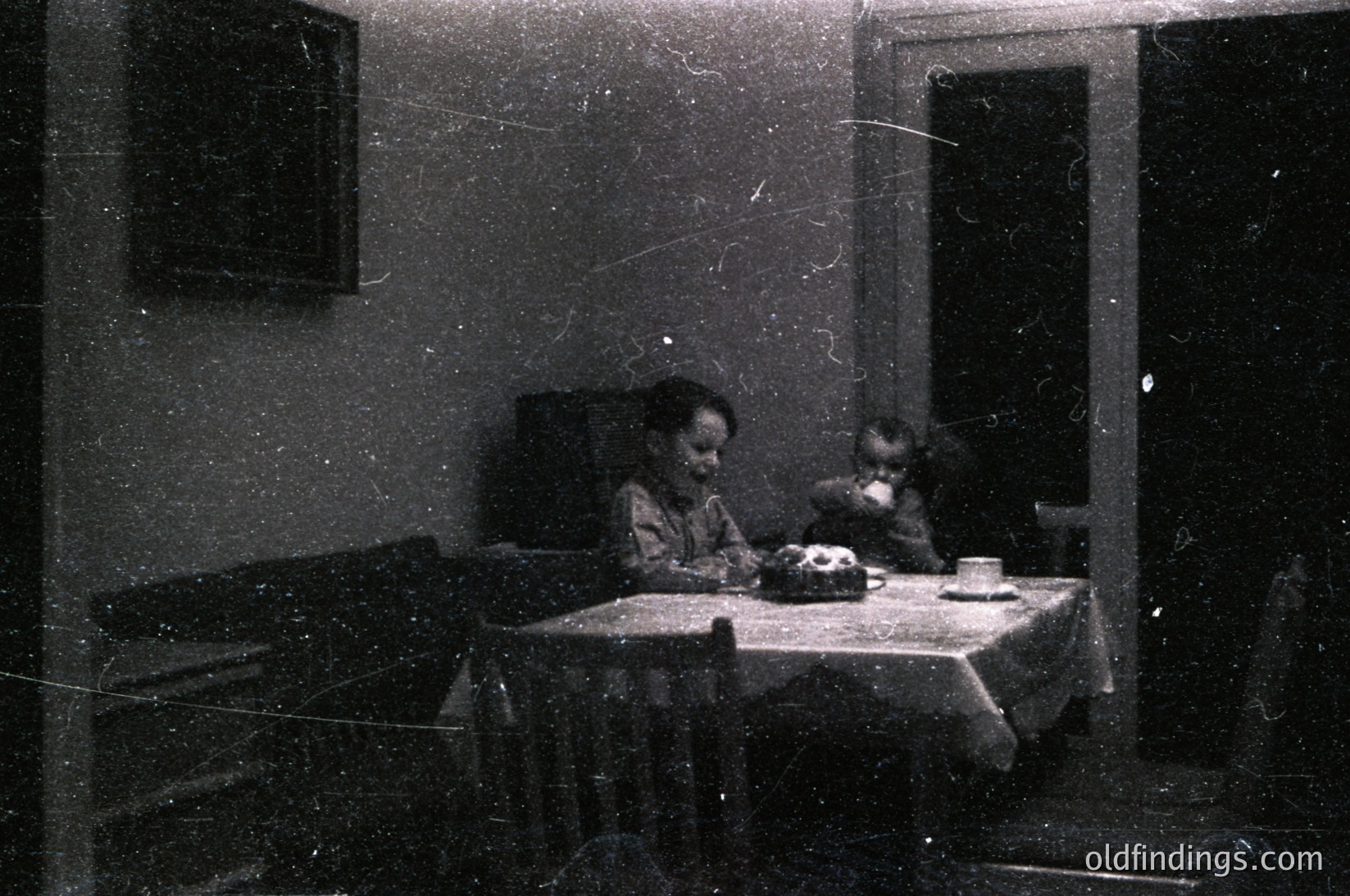 Vintage black-and-white interior shot of two children seated at a small table with a cake, teacup, and cloth napkin. Plain walls, minimalist furnishings, and aged film grain suggest early-to-mid 20th century domestic life.