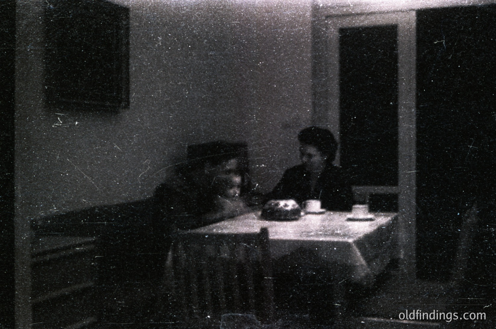 Vintage sepia-toned indoor scene featuring two individuals seated at a small table with a cake and cups. Man in dark cap, woman in high-necked blouse. Simple wooden furniture and framed artwork on walls. Likely mid-20th century domestic setting.