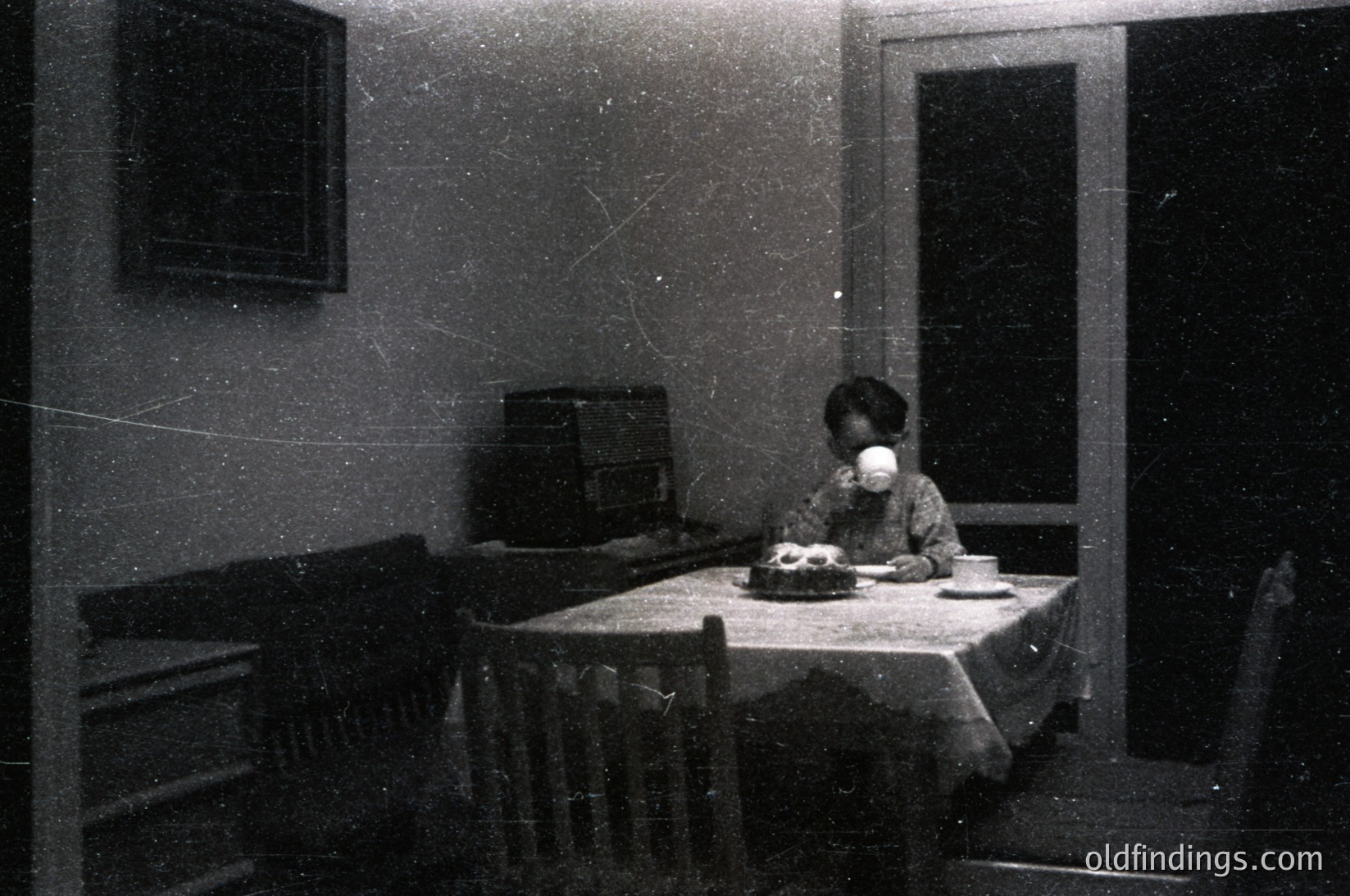 Vintage black-and-white interior shot of a child seated at a round wooden table with a white tablecloth, holding a cup. Sideboard with radio, framed artwork, and wooden chair legs visible. Likely mid-20th century domestic setting. écor