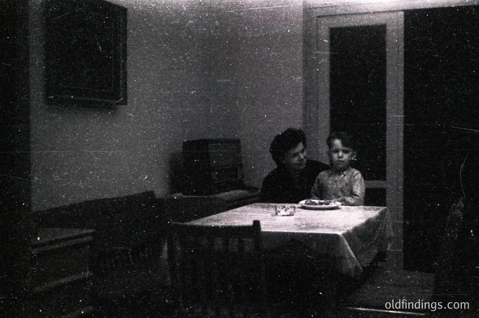 Vintage black-and-white interior shot of a woman and child seated at a small wooden table in a modest room, likely mid-20th century. Tablecloth covers the table, and a framed print hangs above. Simple furnishings include a wooden cabinet and a door leading to another space.