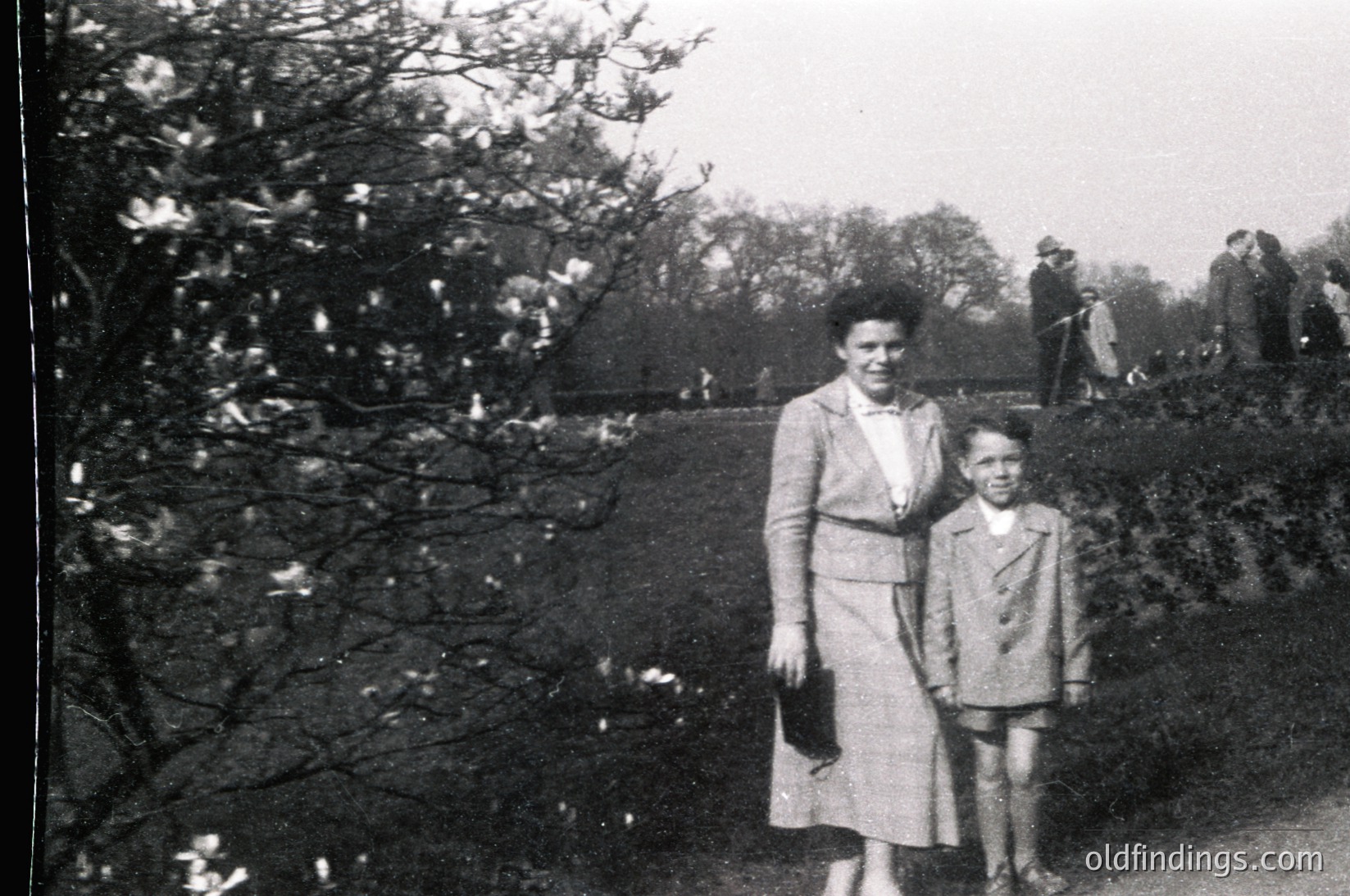 Mid-20th century black-and-white photo of a woman and young boy in a park setting. Woman wears a knee-length dress with a belt and gloves; boy in a short-sleeved jacket and trousers. Leaves and stone wall in foreground.