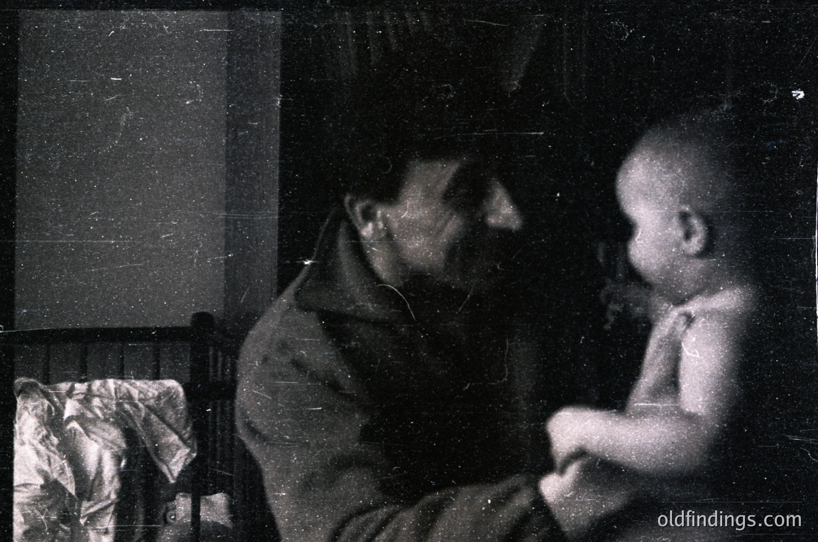 Vintage black-and-white photo of an adult and child sharing an intimate moment indoors. The adult, dressed in a long coat, leans toward the child seated on a bed. A wrapped bundle lies on the bed’s edge. Grainy texture and lighting suggest early 20th-century photography.