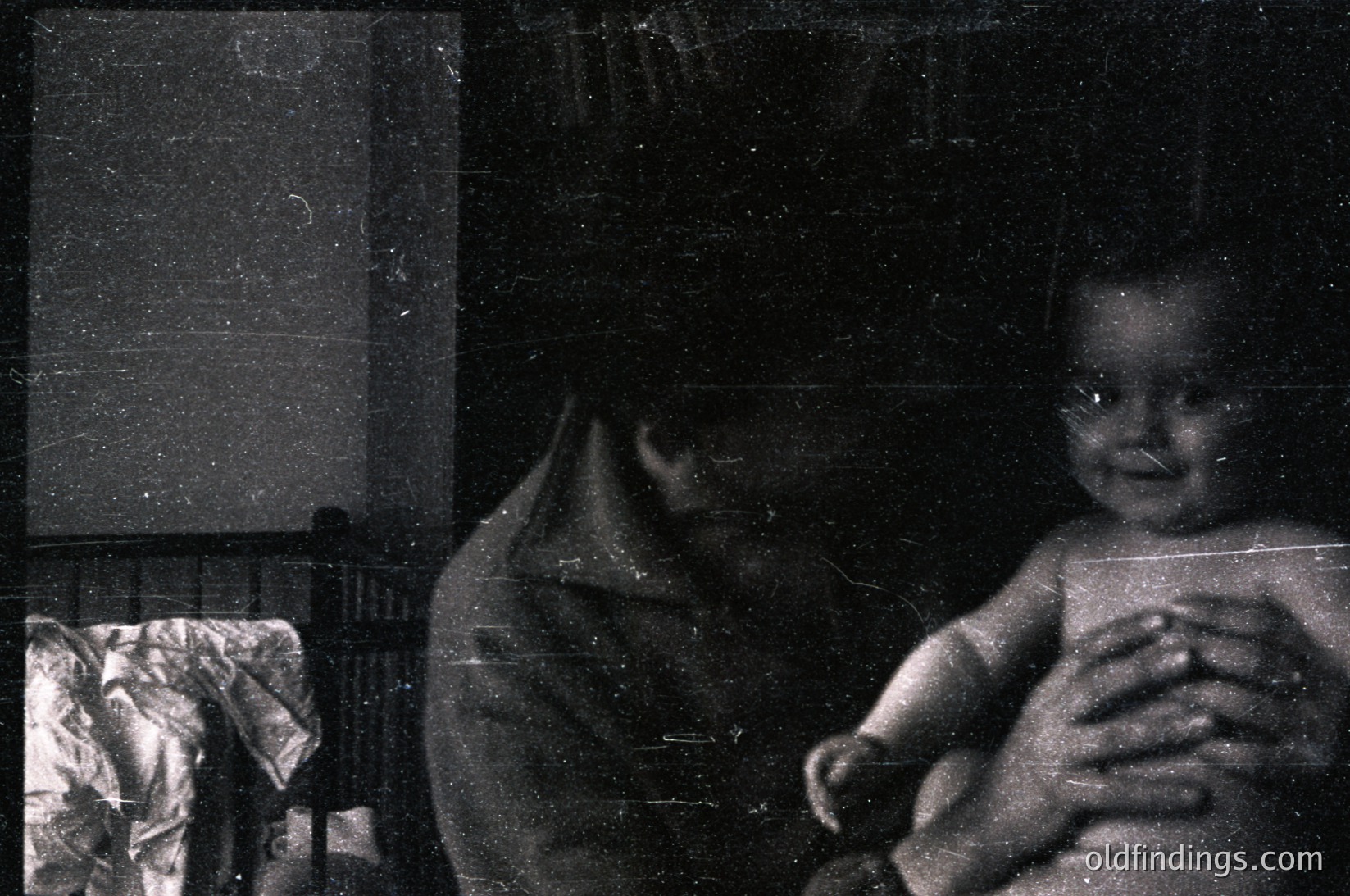 Vintage black-and-white photo of an infant cradled in an adult’s arms, likely mid-20th century. Soft focus and grainy texture suggest film photography. Partial view of a patterned blanket and blurred background furniture. Evokes nostalgic family moments.