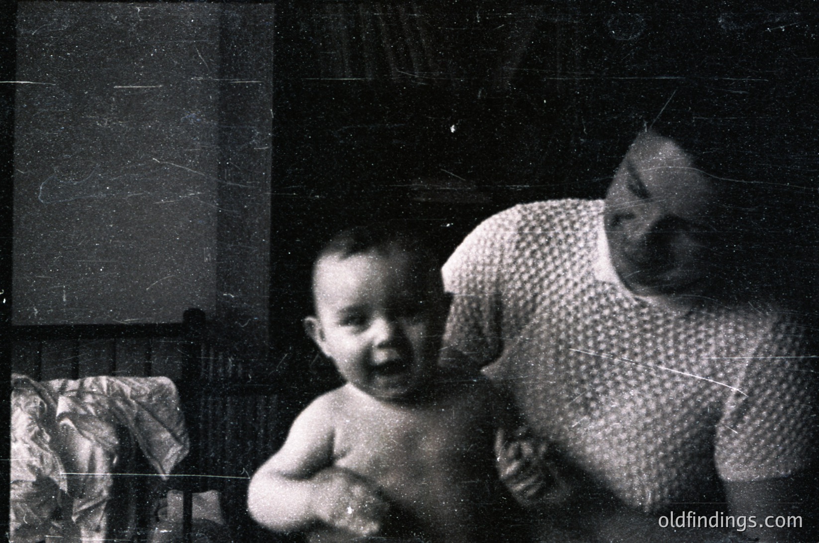 Vintage black-and-white photo of a woman cradling a toddler indoors, likely mid-20th century. The woman wears a patterned blouse, and the child’s bare torso suggests casual, everyday attire. Faded wallpaper and a partially visible armchair indicate a modest residential setting. Emotional warmth and familial bond evident.