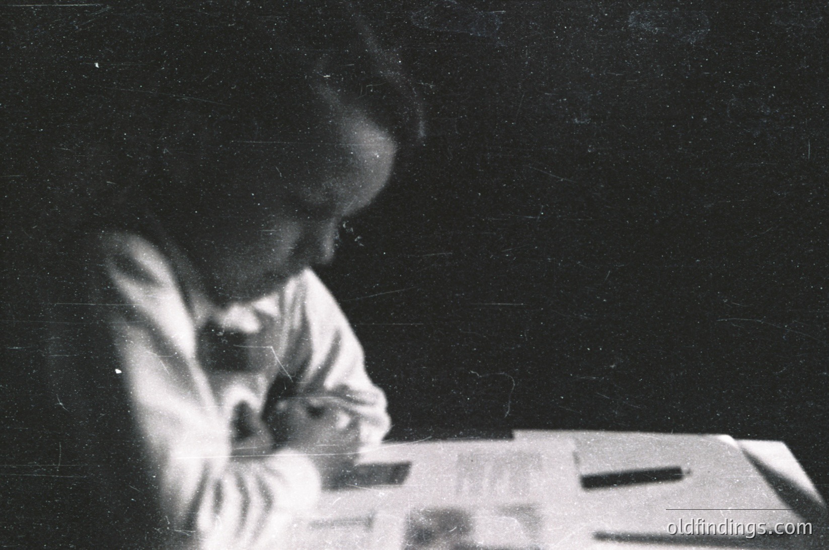 Vintage black-and-white portrait of a seated individual reading or writing at a wooden desk, likely mid-20th century. Soft focus and grainy texture suggest early film photography. Formal attire hints at a formal or educational setting.