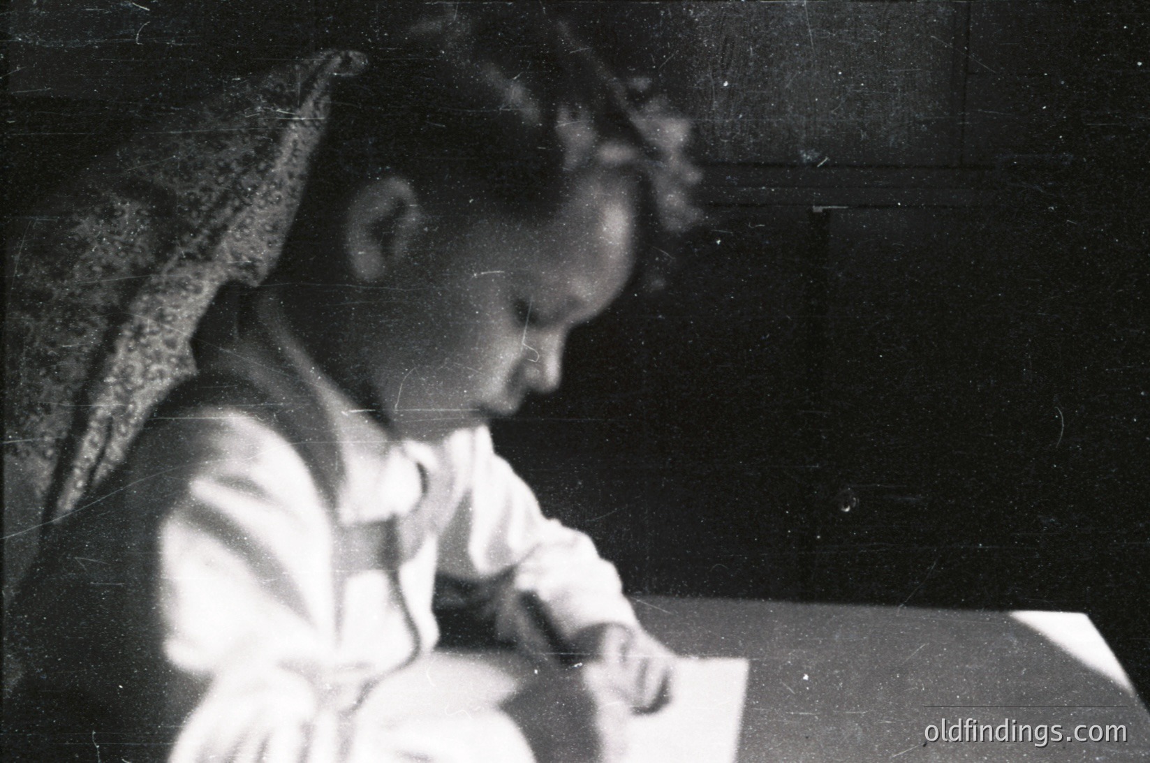 Vintage black-and-white photo of a young child engrossed in writing at a small wooden desk, wearing a white blouse with a lace-trimmed collar. Indoor setting with minimalist furnishings, suggesting early-to-mid 20th century domestic life.