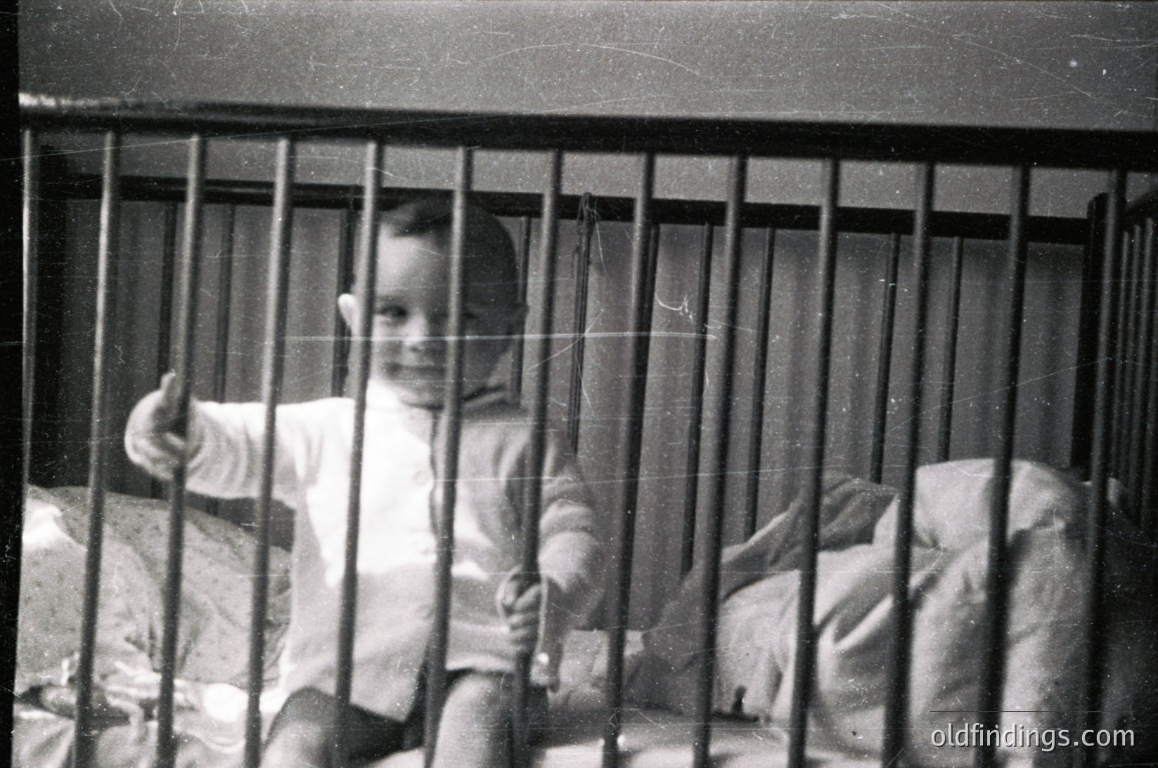 Vintage black-and-white photo of a child in a metal-framed crib, giving a thumbs-up. Simple striped onesie and blanket visible. Likely mid-20th century domestic setting.