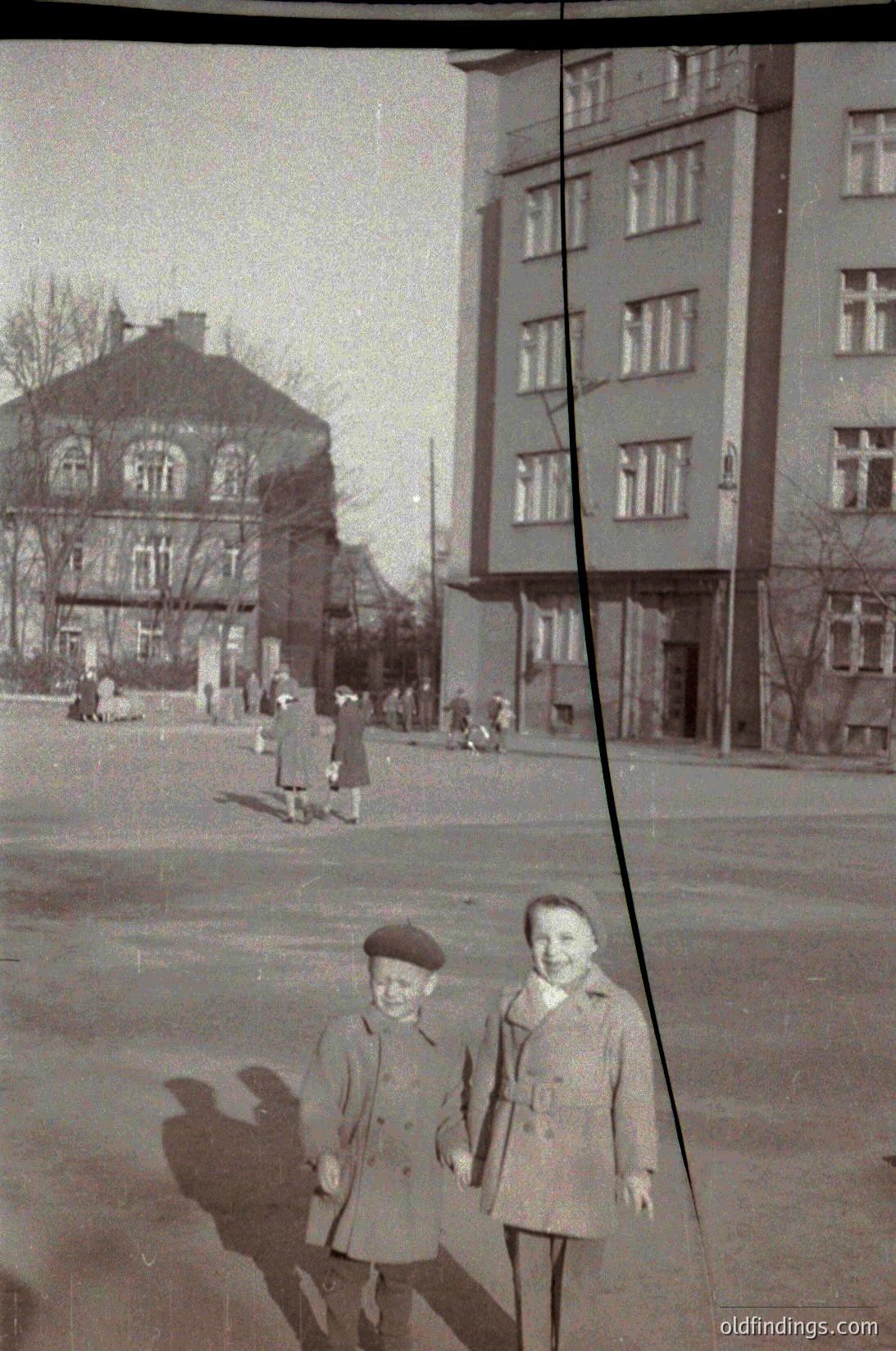 Two children in 1950s-60s Eastern European streetwear pose in front of Soviet-era concrete apartment block. Mixed architecture: older ornate building left, utilitarian block right. Bare trees suggest late autumn/winter. Crowd of people in background adds urban atmosphere.