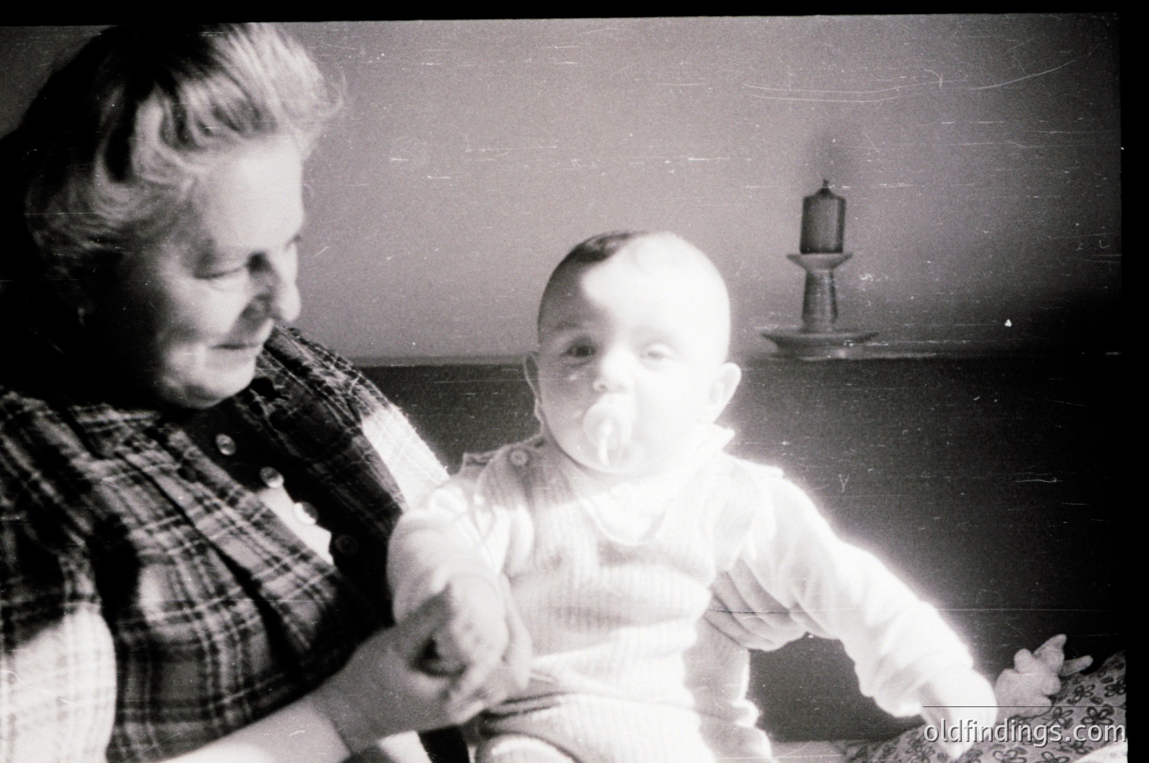 A vintage black-and-white photo of an elderly woman holding a baby indoors, likely mid-20th century. The woman wears a plaid blouse, and the baby sports a white onesie with a pacifier. A lighthouse silhouette decorates the wall behind them, suggesting a seaside or maritime theme.