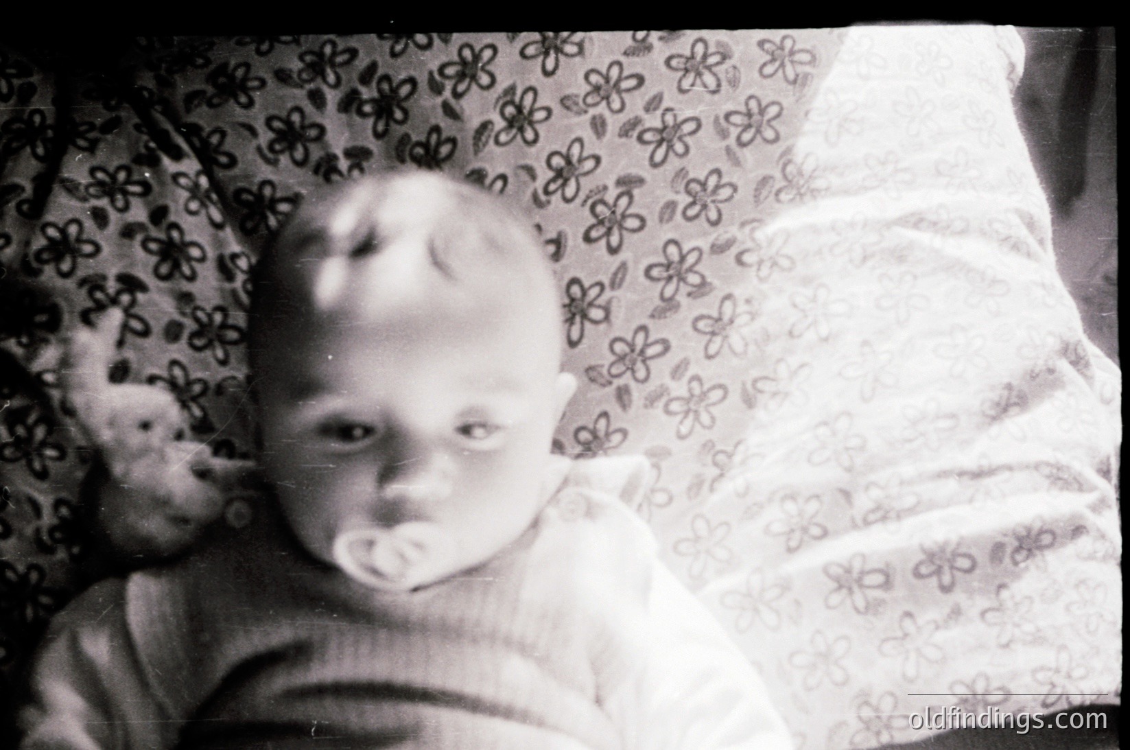 Vintage black-and-white photo of an infant in a floral-patterned blanket, holding a pacifier. Soft focus and grainy texture suggest mid-20th century (1950s–1960s). Classic family portrait style.