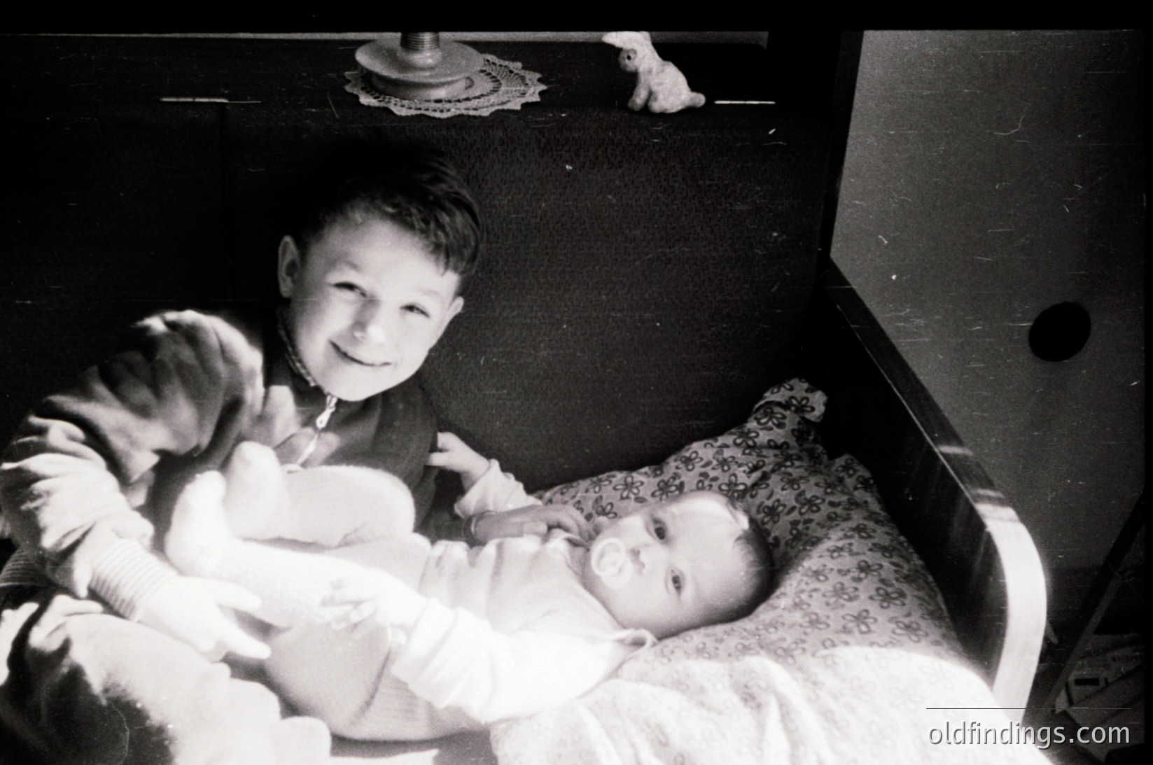Black-and-white snapshot of two boys in a vintage crib, one holding a younger sibling. Mid-20th century domestic setting, likely 1950s–1960s. Distinctive floral-patterned crib sheet and wall-mounted lamp.