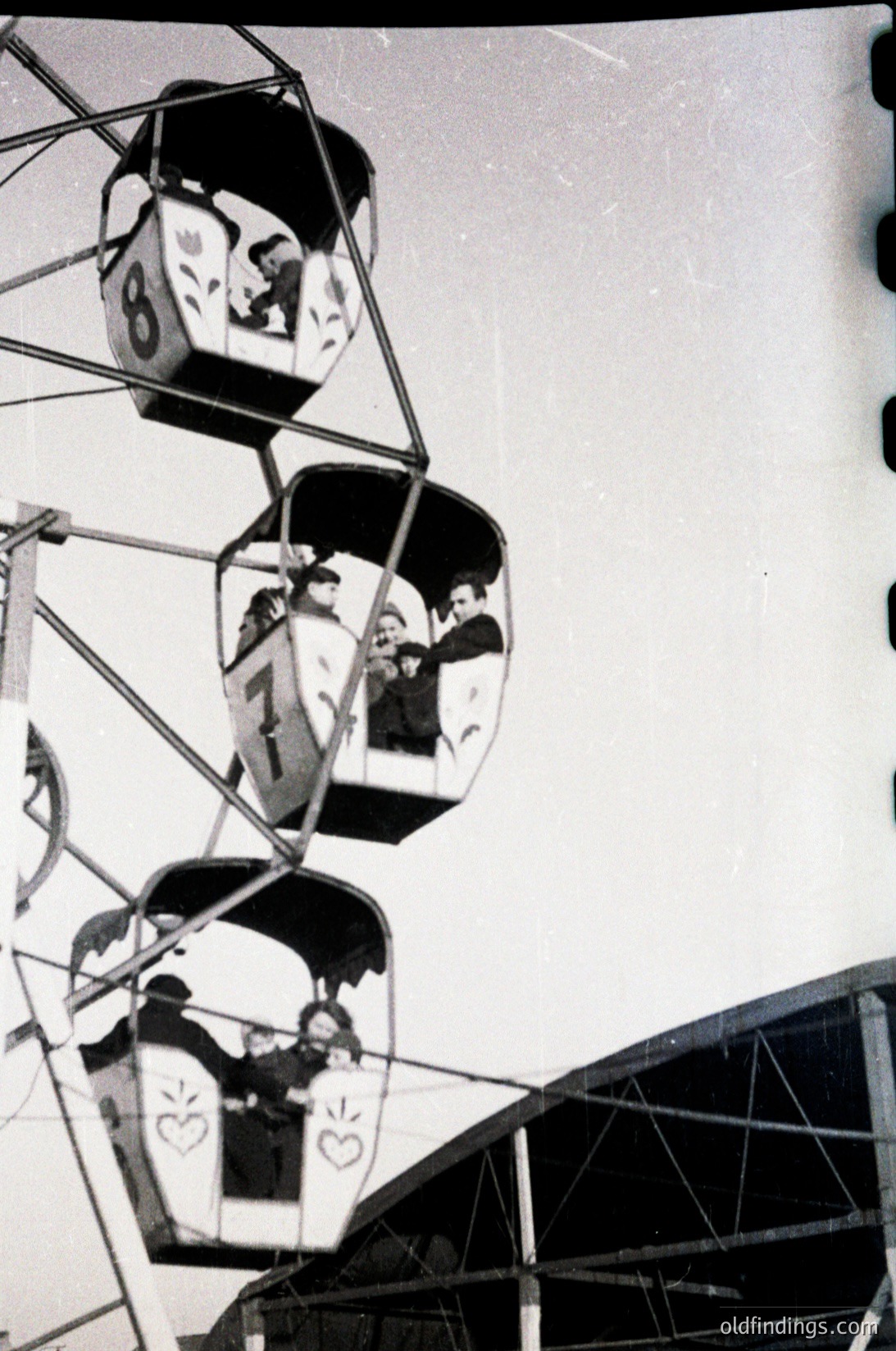 Vintage carousel gondolas with heart-shaped logos, mid-20th century design. Black-and-white photo captures families enjoying rides, suggesting a mid-century amusement park or fairground setting. Distinctive retro aesthetic ideal for historical research or nostalgic design references.
