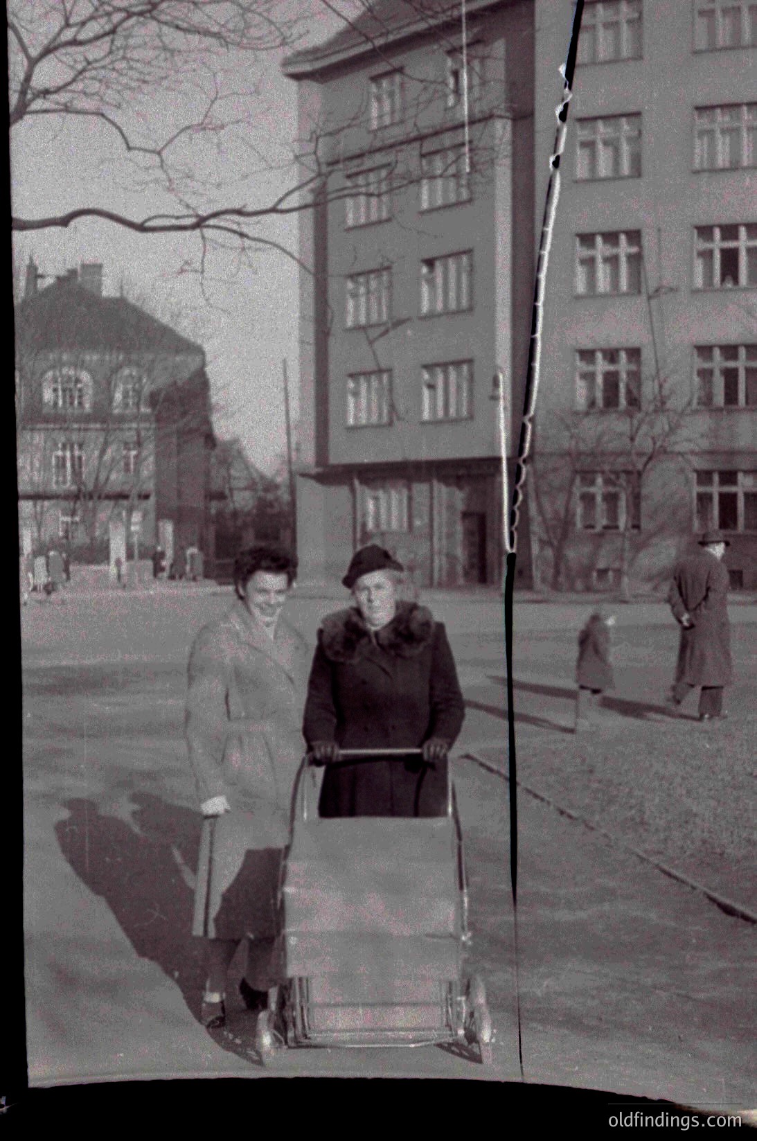 Two women in 1950s-era clothing pose with a baby carriage on a paved urban street flanked by mid-rise brick buildings. One wears a hat and coat; the other holds a cane. Snow patches and bare trees suggest winter.