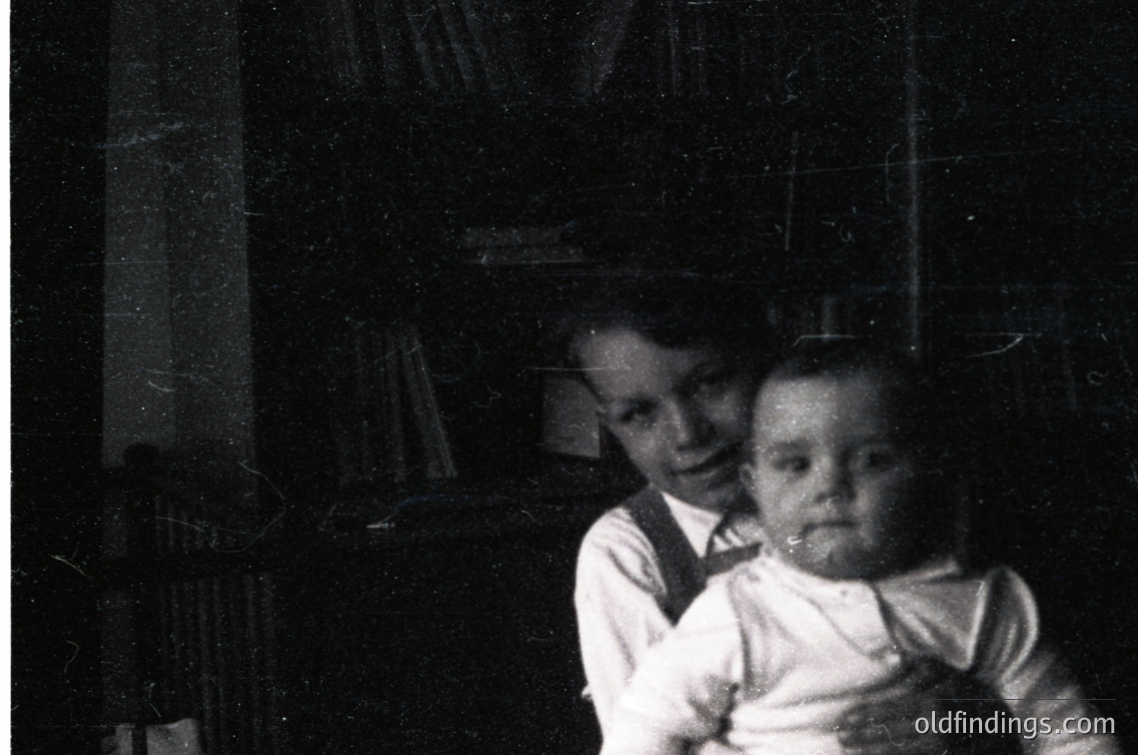 Vintage black-and-white portrait of an older child holding a younger sibling indoors, likely mid-20th century. Bookshelves with worn volumes and a dark, textured wall in background. Formal attire suggests a posed family moment.