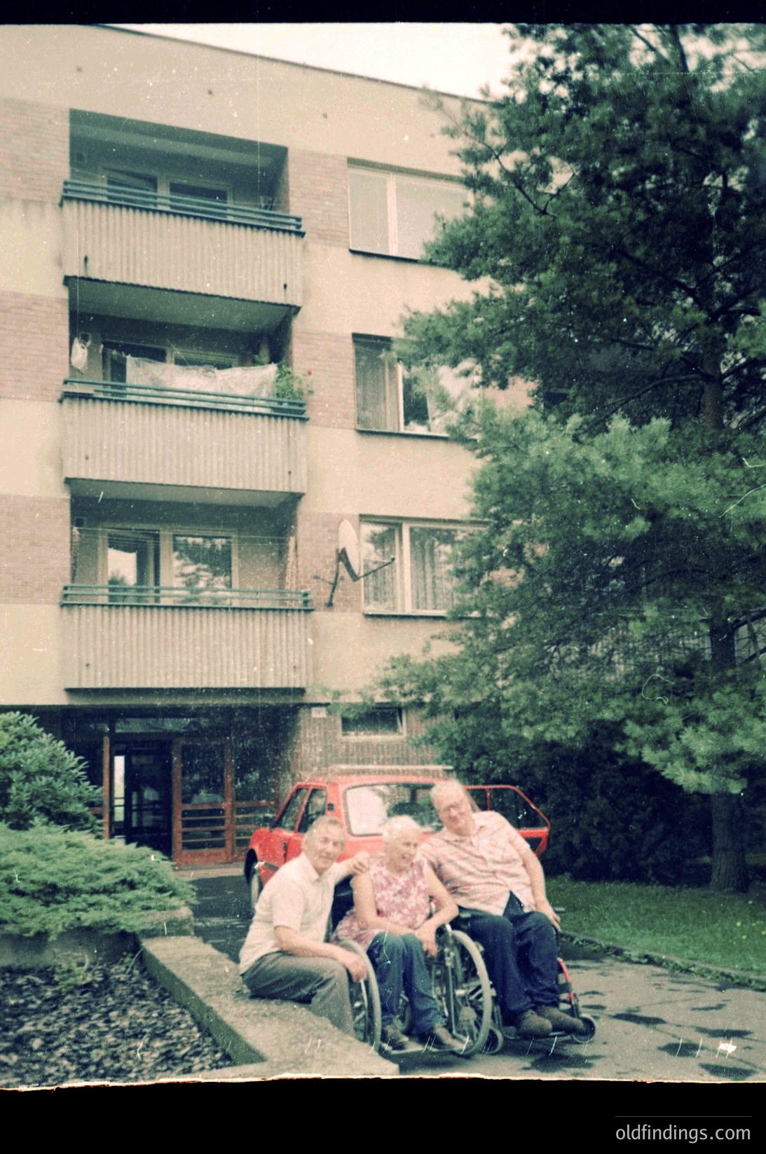 Vintage 1970s-era apartment block with concrete balconies, featuring three individuals—two seated, one in a wheelchair—posing in front of a red car. Urban residential setting with greenery and paved walkway.