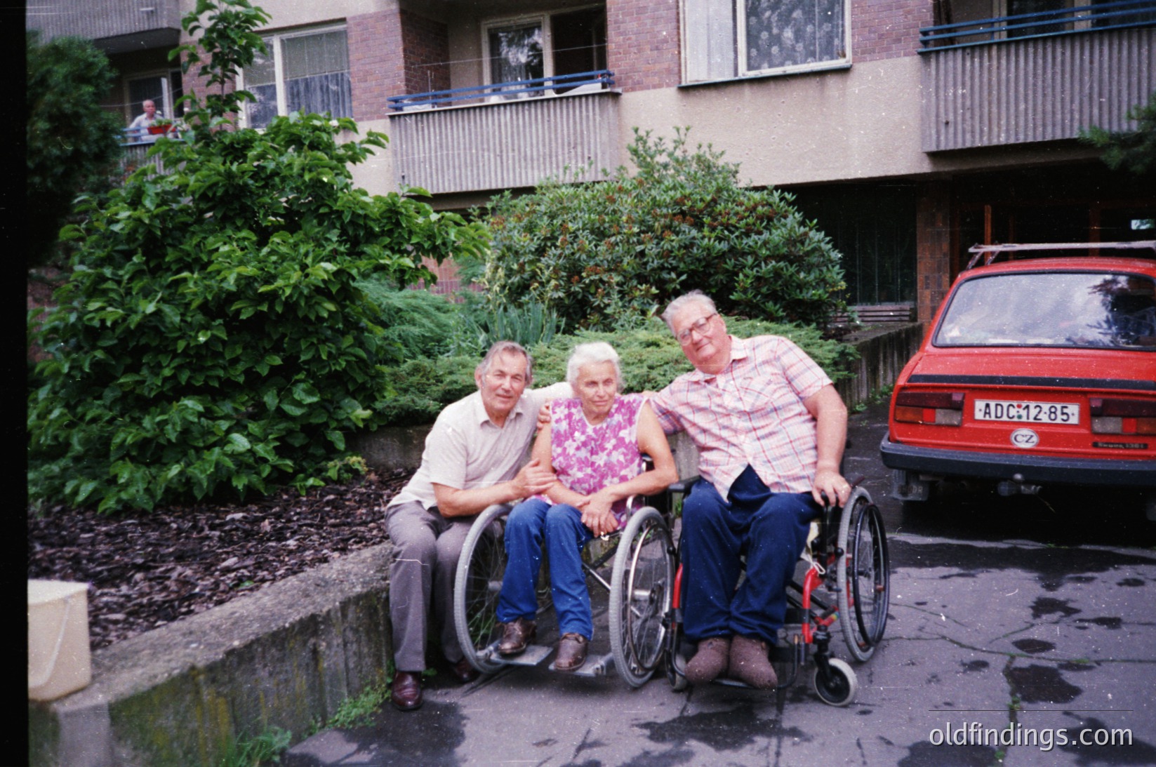 Three adults in wheelchairs pose outdoors near a multi-story residential building with balconies, likely from the 1980s–1990s. The man on the left wears a light shirt and dark pants; the woman in the center wears a floral blouse; the man on the right wears a striped shirt and dark pants. A red car with a visible license plate (partially obscured) is parked behind them. Lush greenery and a paved pathway frame the scene.