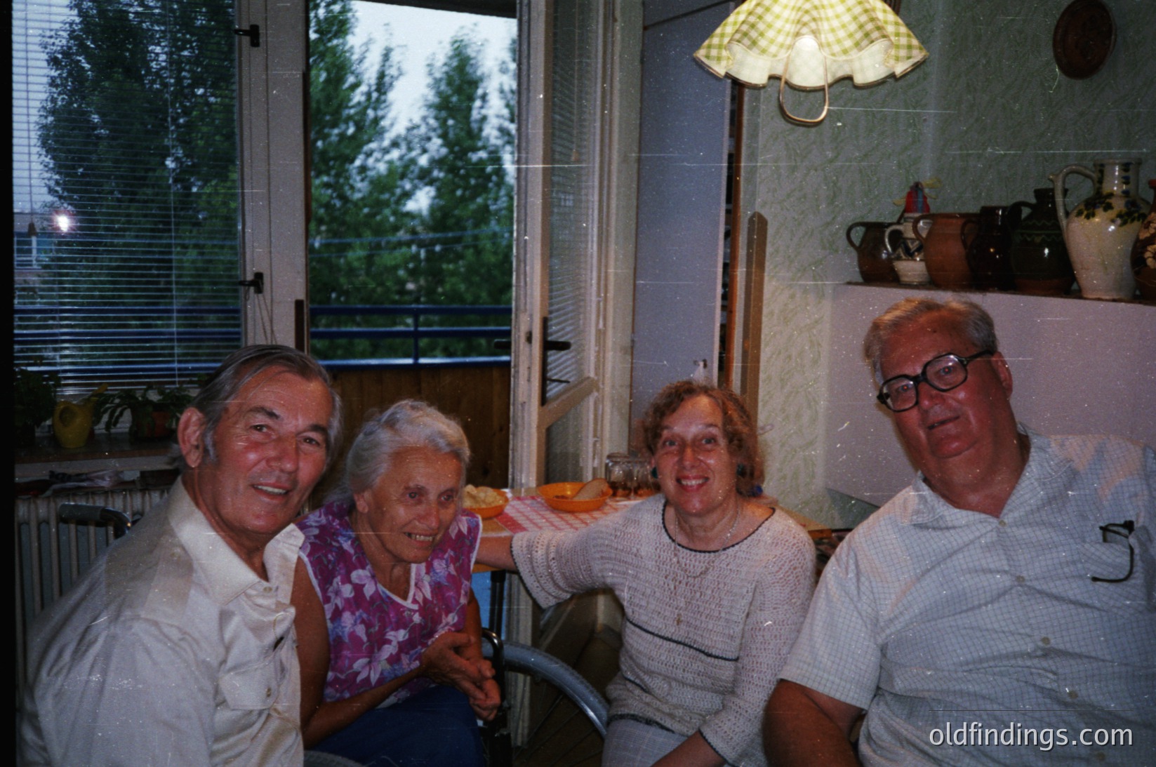 Four adults pose indoors near a window showing greenery, likely a balcony or terrace. Mid-20th century home décor: ceramic jars, a yellow checkered umbrella, and a white radiator. Clothing suggests late 1970s–early 1980s European style.