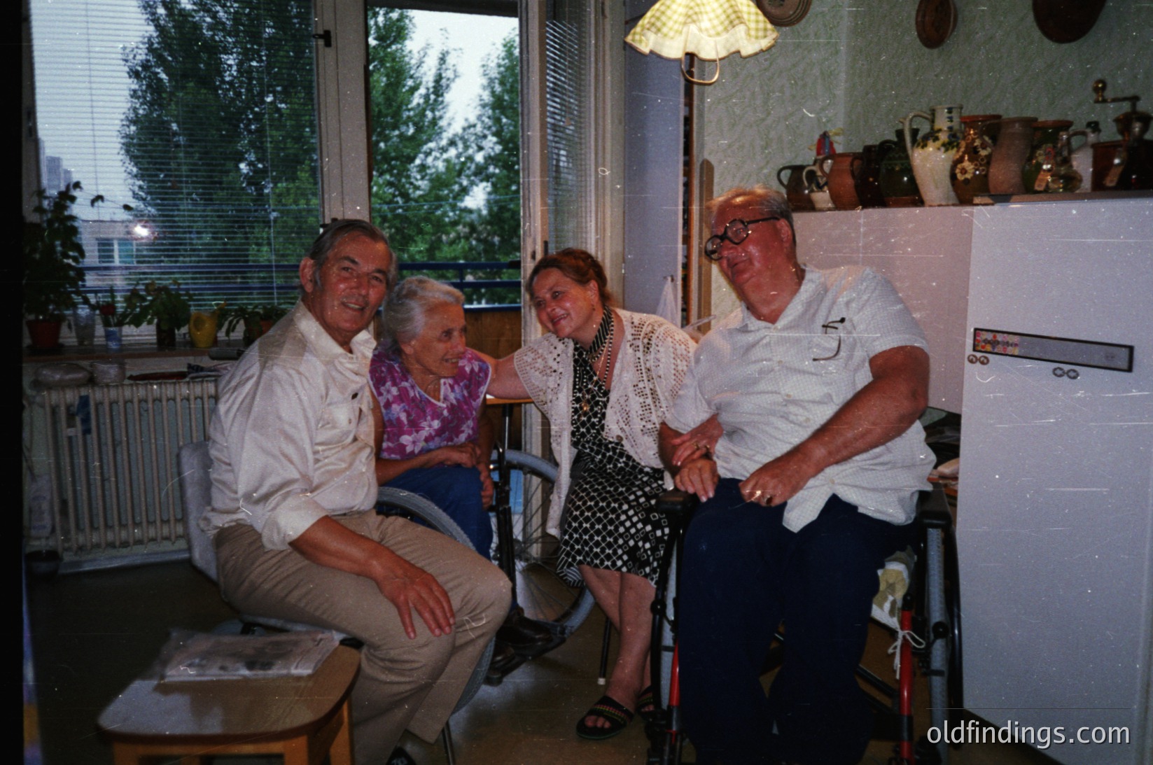 Four adults pose indoors in a mid-century home, likely Eastern Europe, 1970s–1980s. A man and woman in wheelchairs sit beside a standing woman in a floral dress, with another man leaning on a cane. Decor includes a vintage fridge, ceramic jars, and a lamp. Warm, lived-in atmosphere.