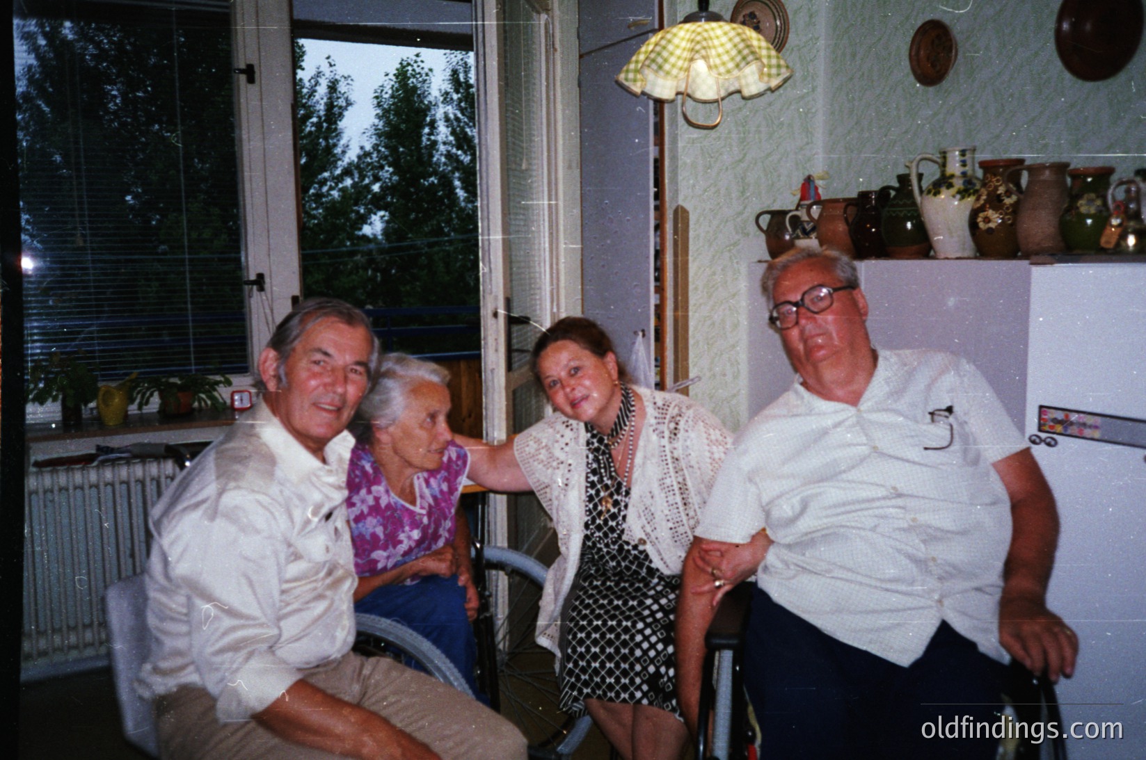 Four adults pose indoors, likely mid-1980s–1990s Eastern Europe. A man and woman in wheelchairs sit beside two standing individuals in a cozy room with floral wallpaper, ceramic decor, and a vintage umbrella lamp. Warm lighting and a view of greenery outside suggest a domestic, communal moment.