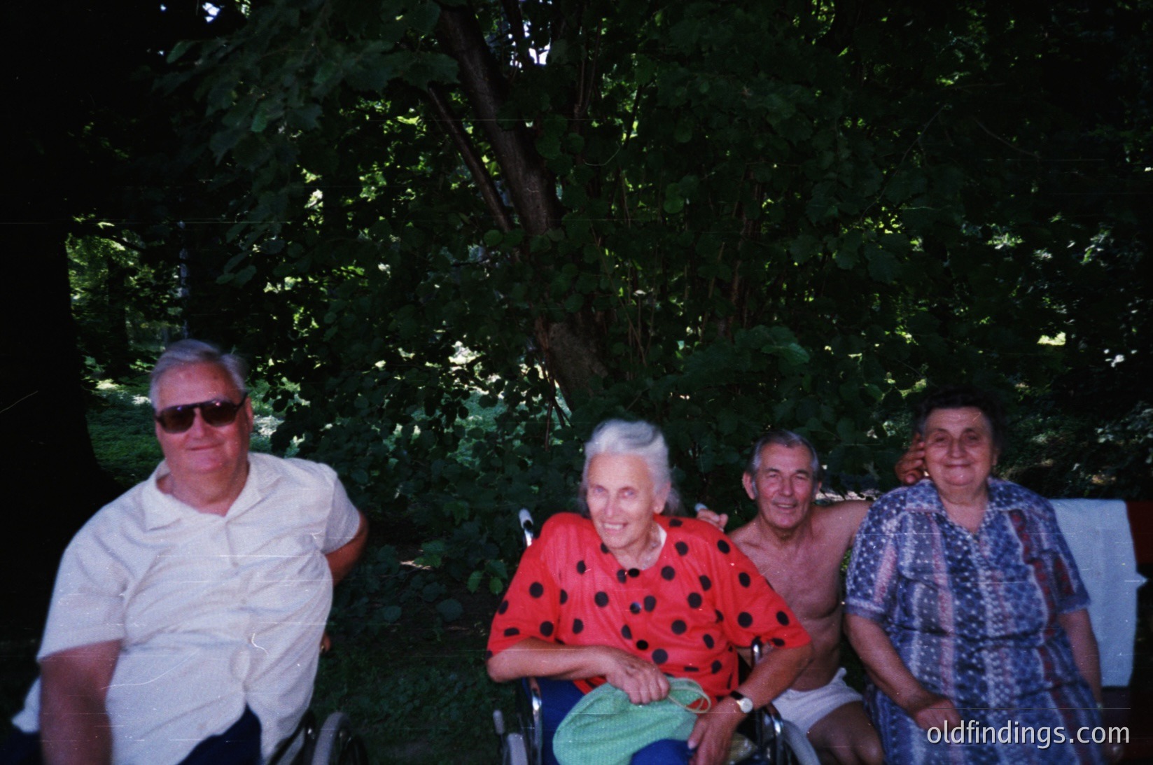 Four adults pose outdoors under dense foliage, likely in a garden or park setting. The man on the left wears sunglasses and a white polo shirt; the seated woman in the center dons a polka-dotted red dress. The man beside her wears a white sleeveless shirt, and the woman on the right wears a patterned blouse. Their attire suggests a mid-20th century timeframe (, ). The lush greenery and relaxed poses evoke a nostalgic, communal atmosphere.