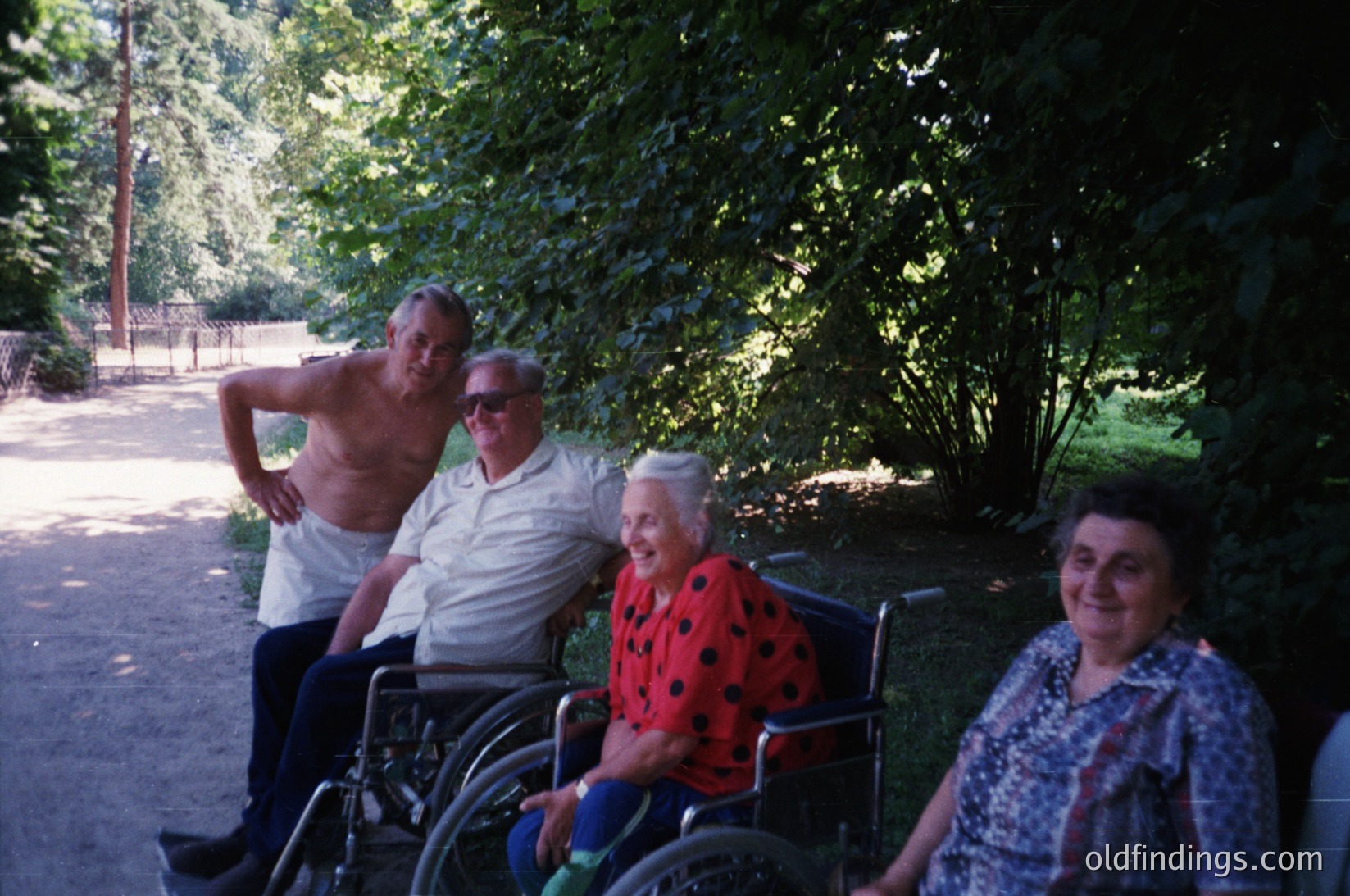 Four seniors pose outdoors in a shaded park setting, likely mid-20th century. Two men stand—one shirtless, one in a light-colored shirt—while two women sit in wheelchairs, one in a red polka-dot dress, the other in patterned clothing. Lush greenery and a paved path frame the scene.