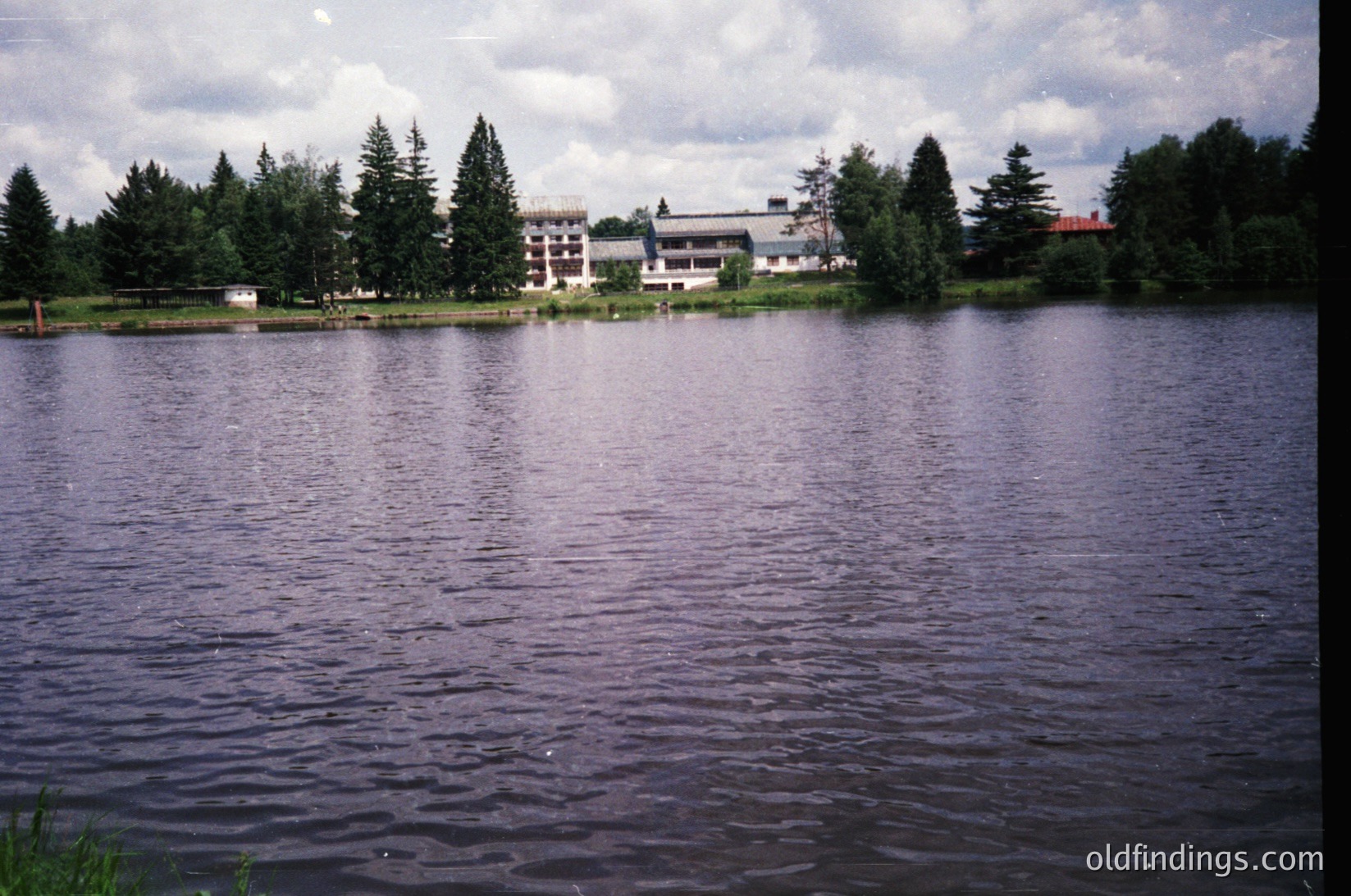 Lakefront view of a mid-20th century resort complex with modernist architecture. Low-rise buildings with flat roofs and large windows flank a forested shoreline. Calm water reflects muted greens and grays under overcast skies.