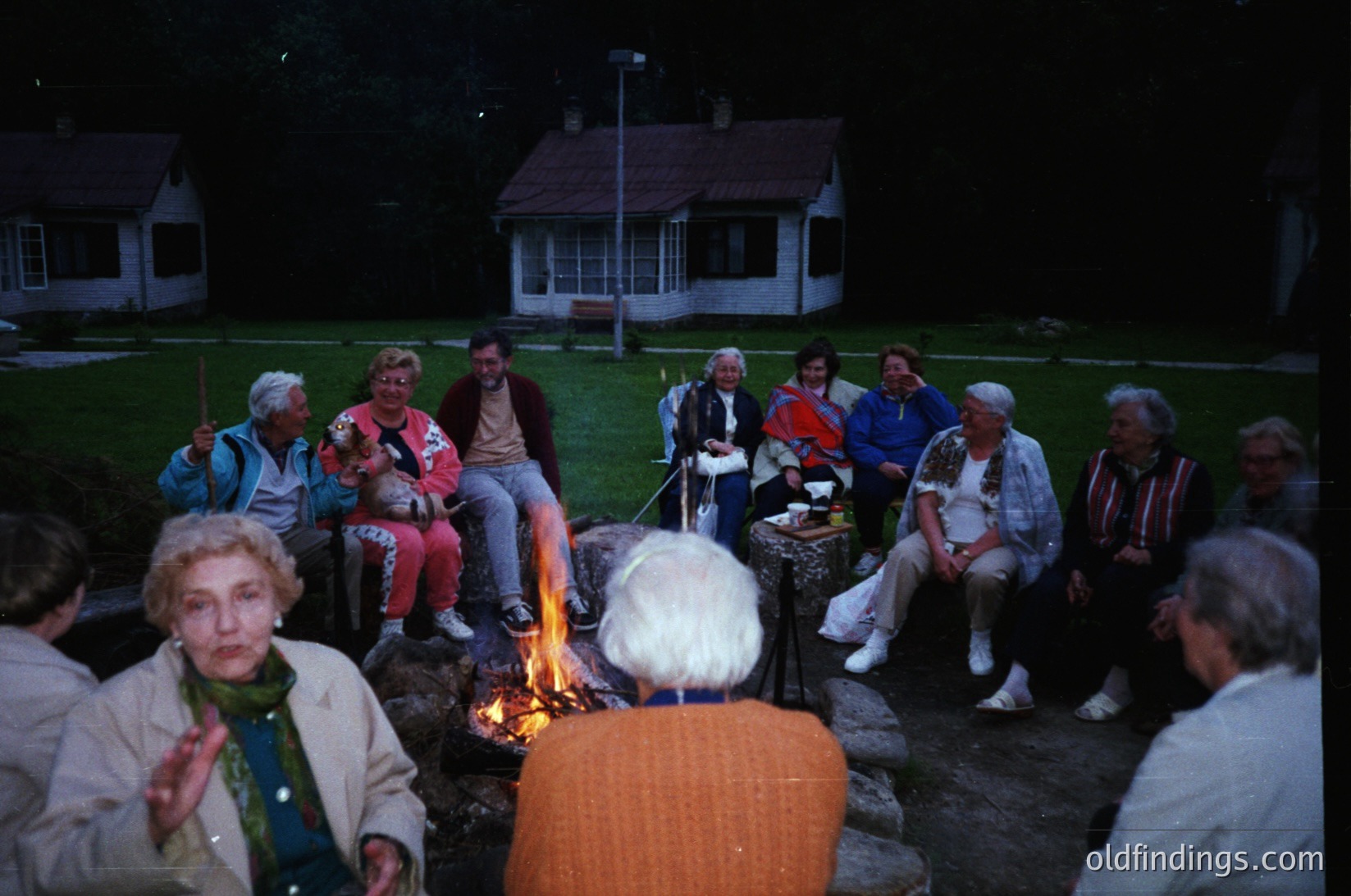 Group of 12+ adults gathered around an outdoor fire pit in a residential backyard, 1970s-1980s. Casual attire includes sweaters, scarves, and retro patterns. Suburban setting with single-story homes and well-manicured lawns. Warm, intimate evening gathering.
