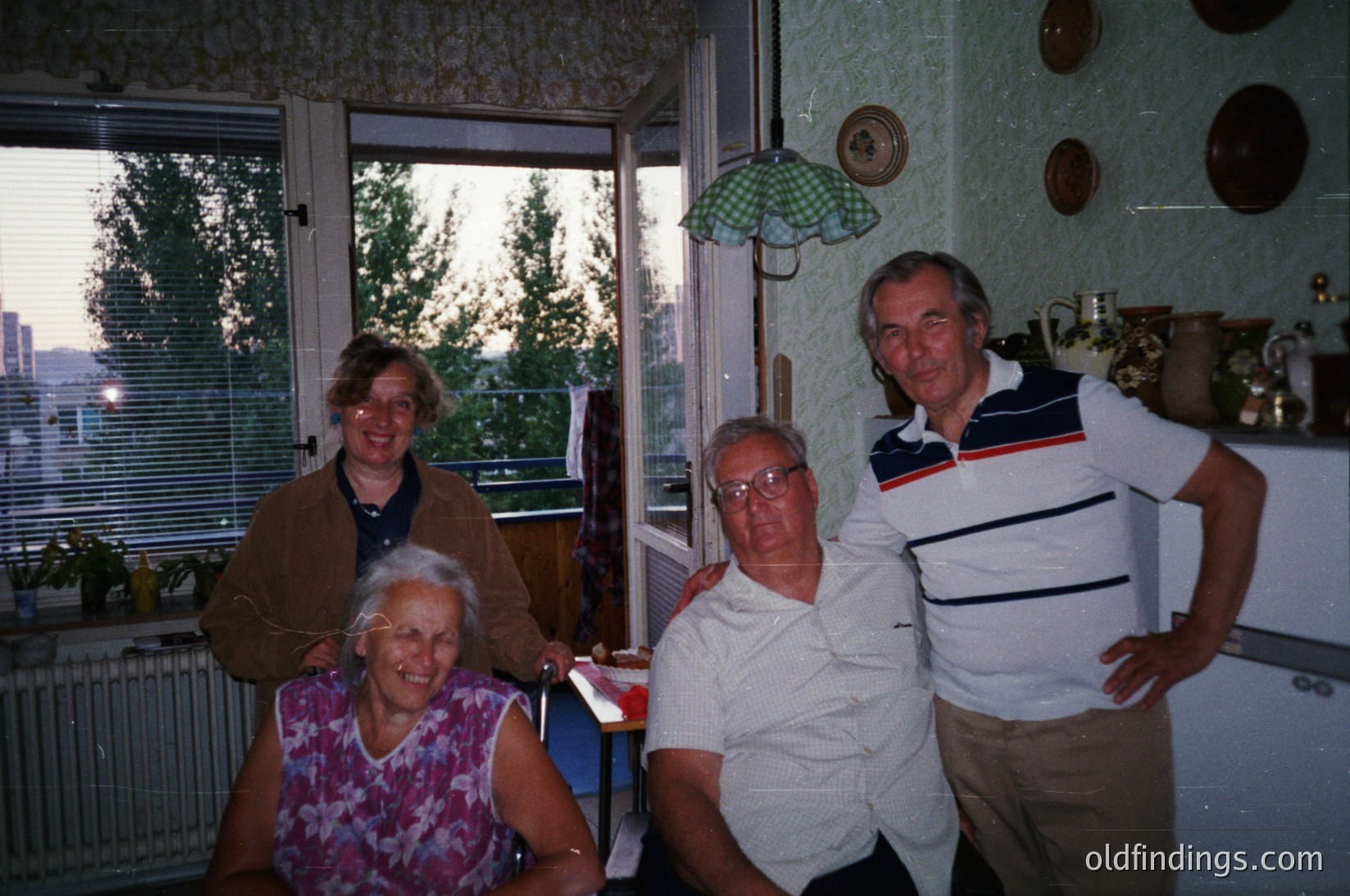 Four adults pose indoors in a mid-century kitchen, likely 1970s–1980s. Light-colored walls, checkered curtains, and ceramic plates on display. Men wear striped polo shirts; women in floral blouses. Window reveals urban skyline.
