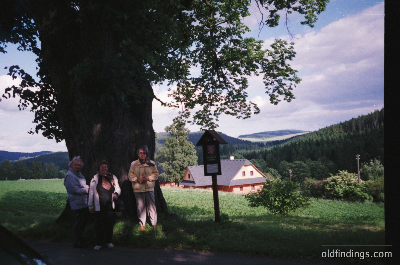 Three individuals pose under a large tree beside a vintage signpost in a rural landscape. The signpost displays a red-and-white directional marker. A red-roofed house with white trim sits in the background, surrounded by green fields and forested hills under a partly cloudy sky.