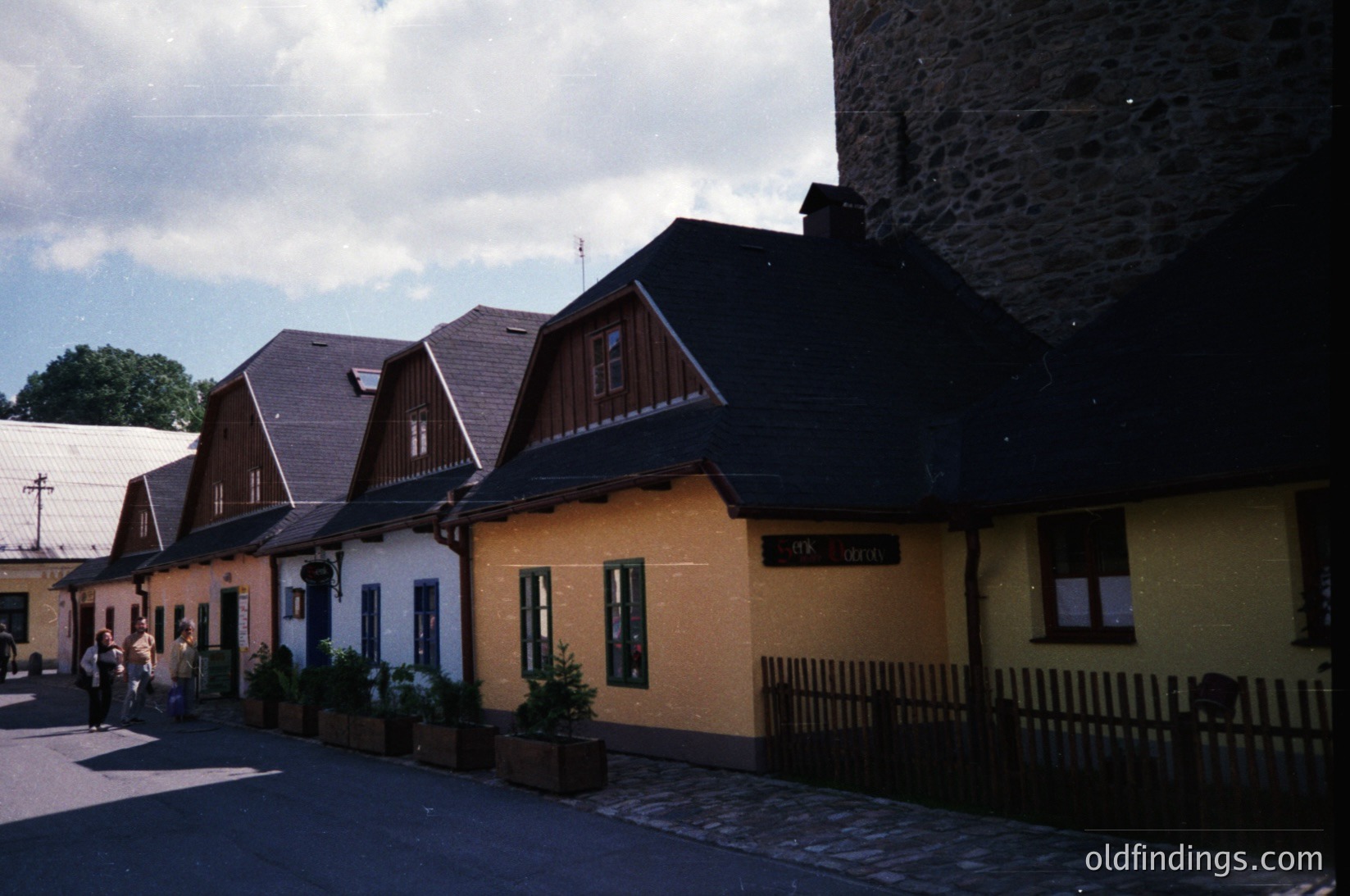 Colorful row of 1960s-era European village houses with steep gabled roofs, pastel facades (, , ), and potted plants. Cobblestone street and stone tower in background suggest historic European architecture. Ideal for retro design references or cultural studies.