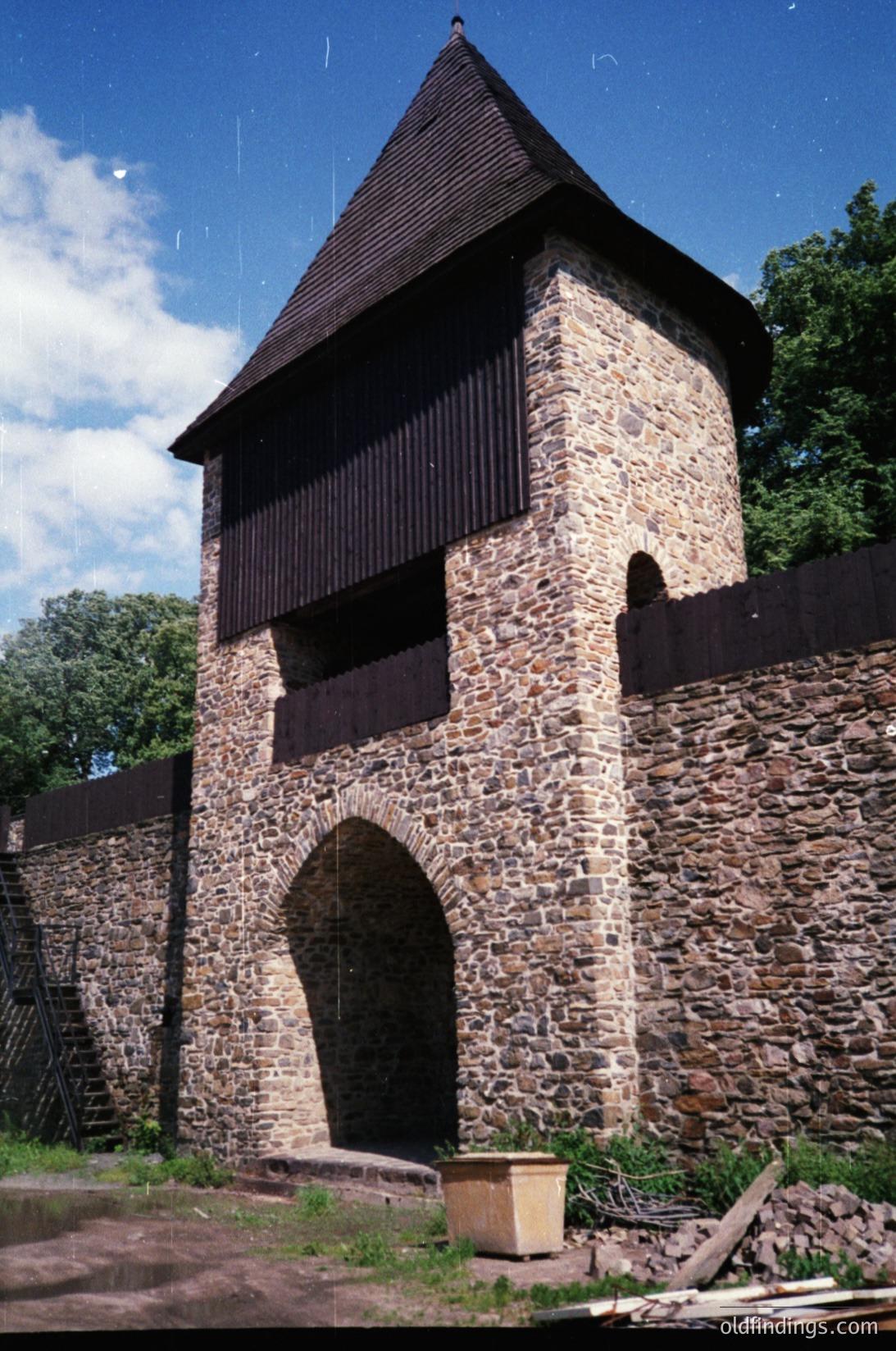 Historic stone watchtower with timber-framed upper section, featuring a steep gable roof and arched entrance. Constructed from irregularly cut stones, likely medieval or early modern defensive architecture. Surrounded by a low stone wall with a wooden barrel chest resting beside the entrance. Lush greenery and clear blue sky in background.